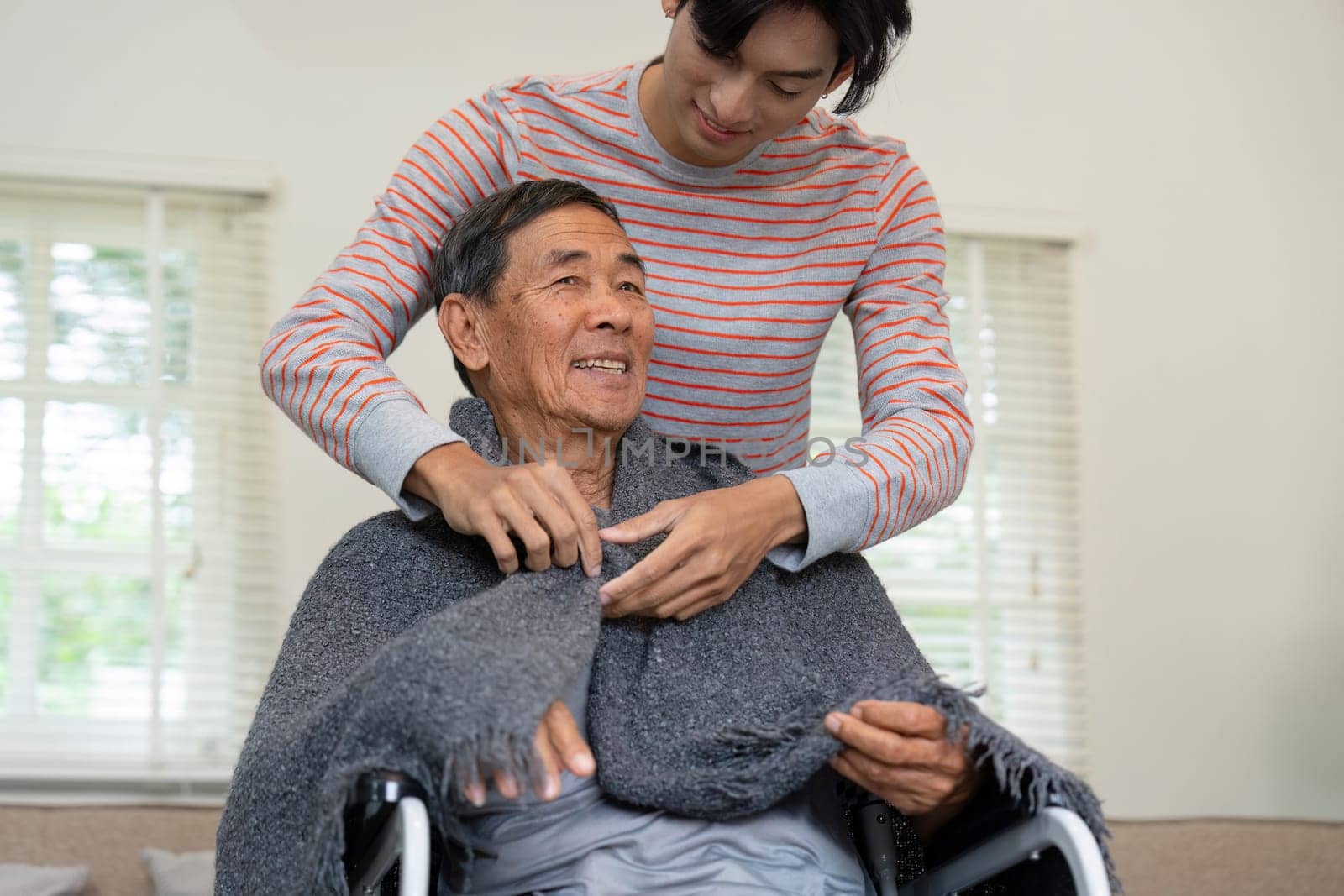 A son assists his elderly father in adjusting a blanket while he sits in a wheelchair, emphasizing care and family support in a comforting home atmosphere.