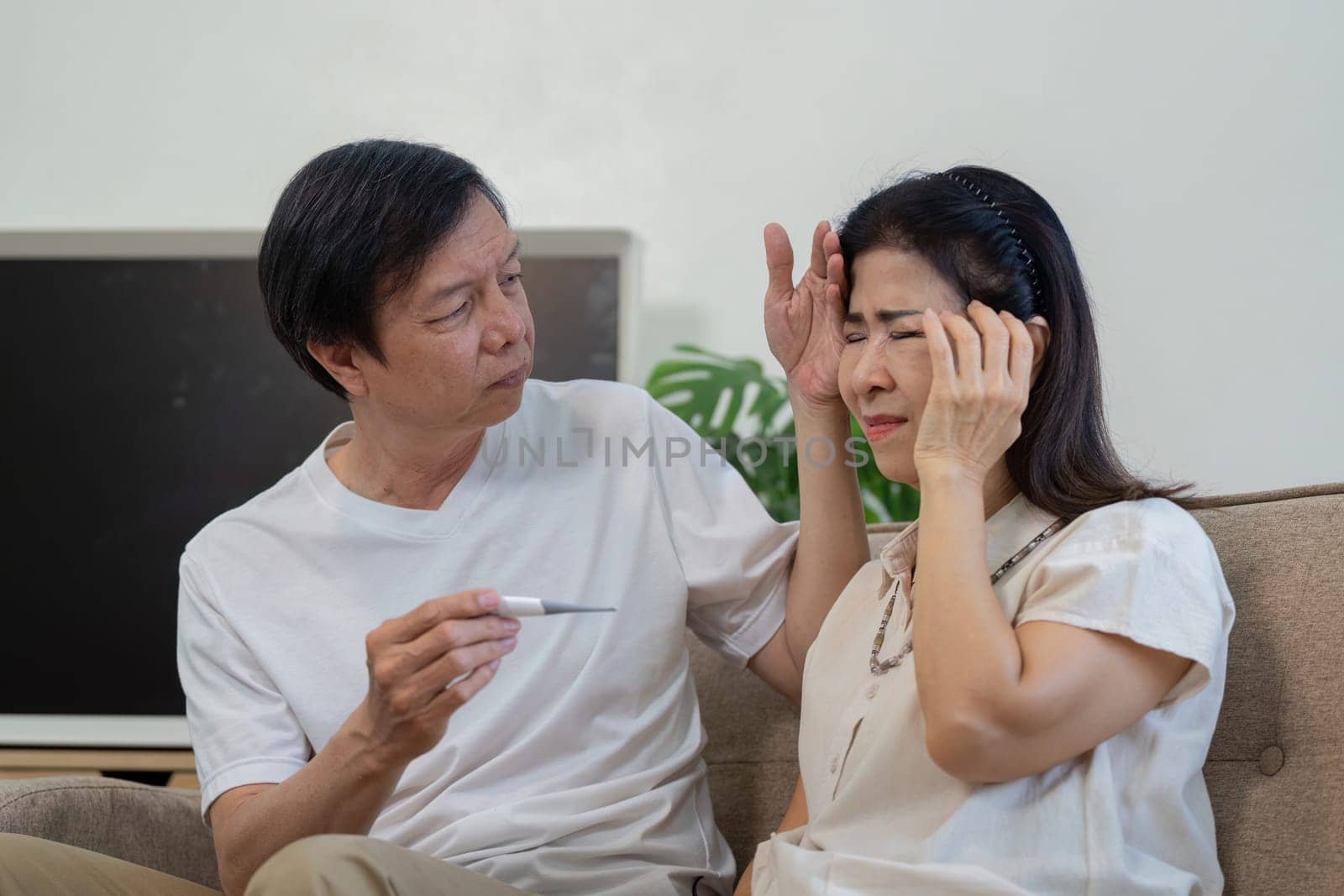 An elderly couple at home, with one partner holding a thermometer, concerned for the other who is suffering from a headache.