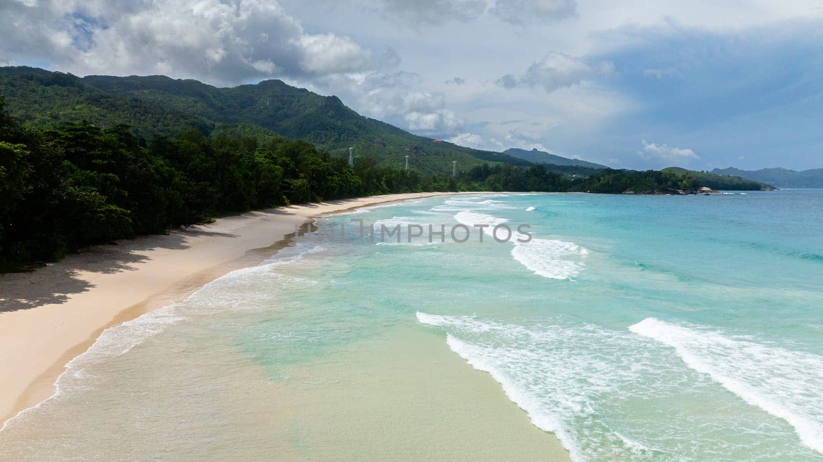 White sandy beach meets turquoise waves, surrounded by tropical vegetation and greenery. Seychelles, Mahe. Grand Anse Beach.