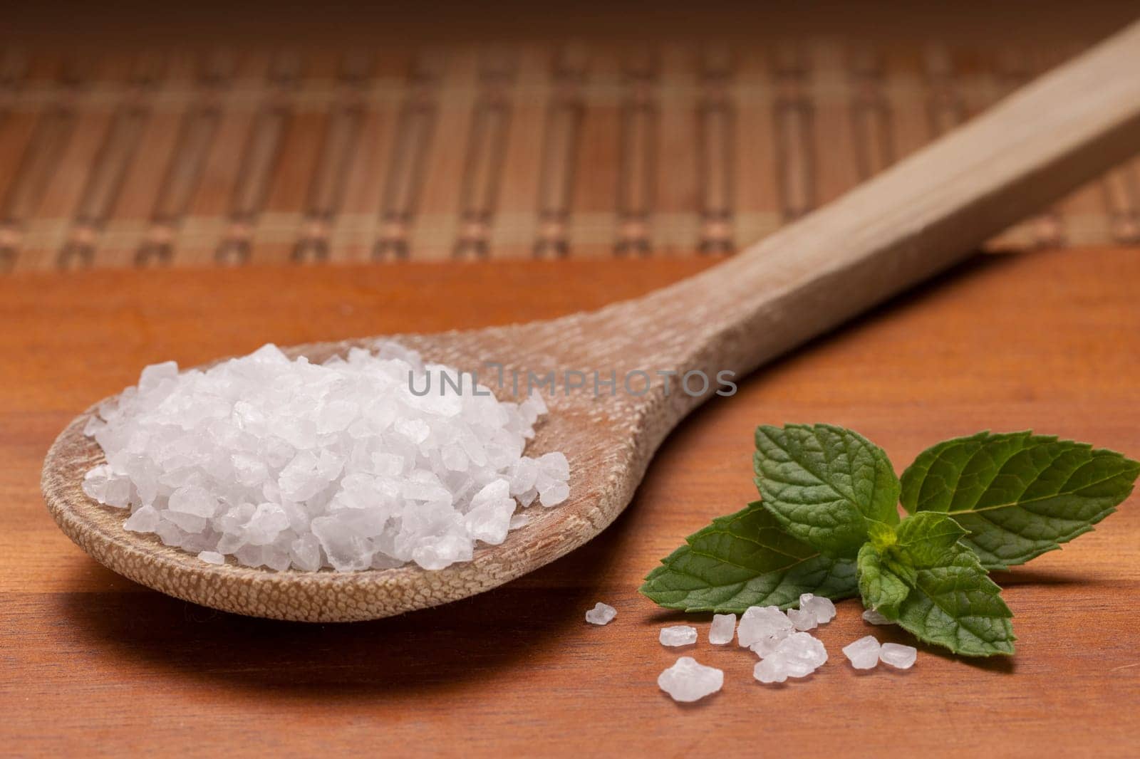 A close up image of a wooden spoon filled with sea salt and a green leaf on a cutting board.