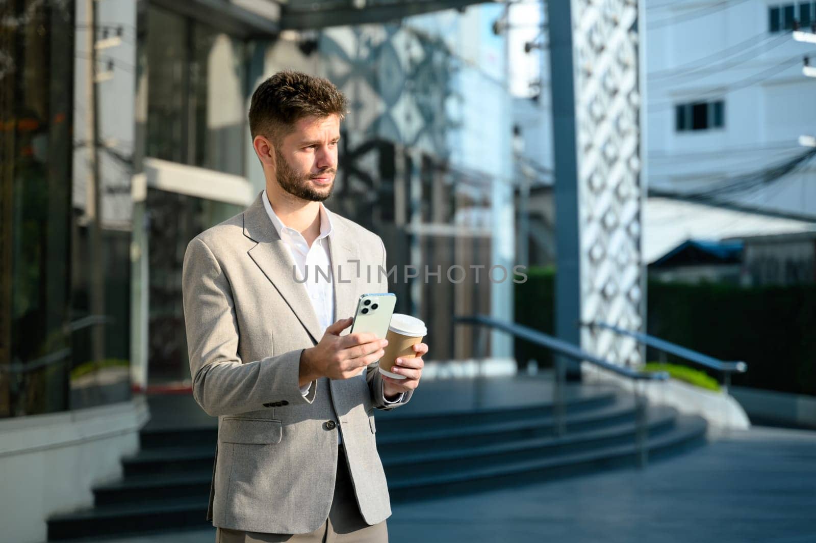 Young businessman in a gray suit holding smartphone standing outside a modern office building.