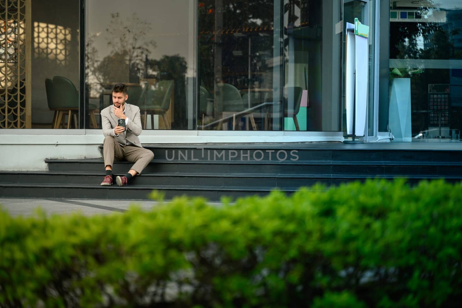 Young professional in suit checking smartphone sitting outdoors at office building.