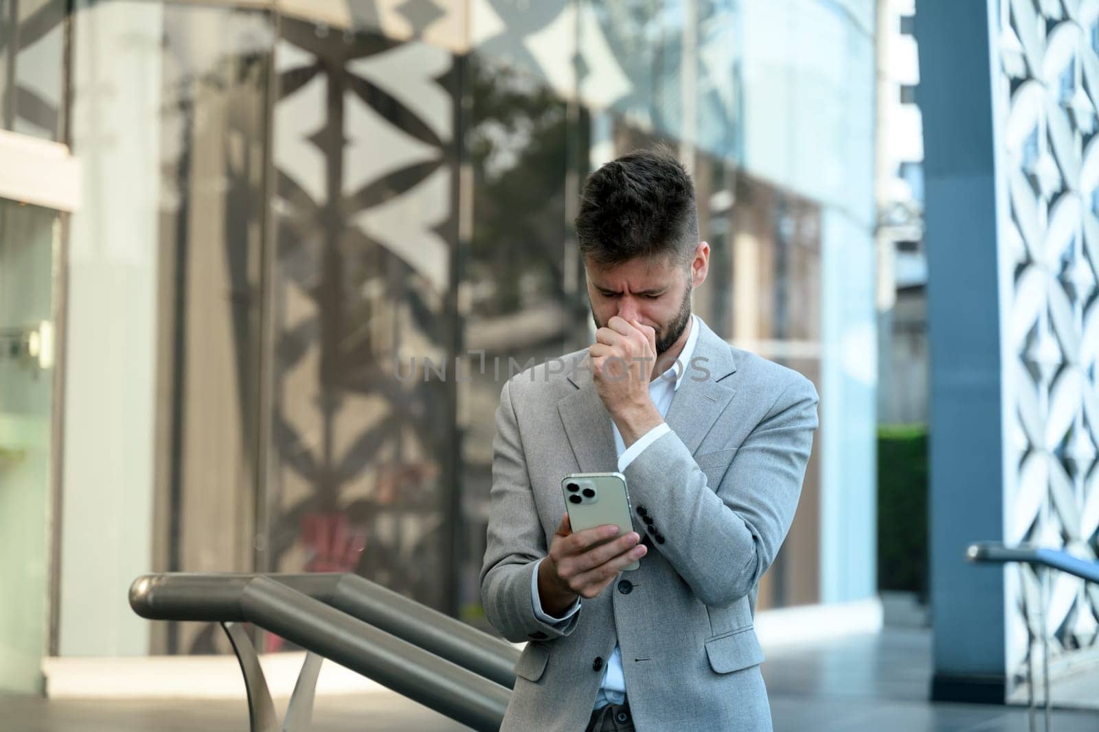 Stressed businessman reacting to bad news on smartphone, standing outside office building.