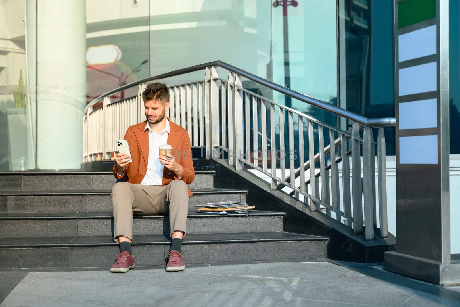 Young businessman sitting on outdoor office stairs and checking mobile phone.