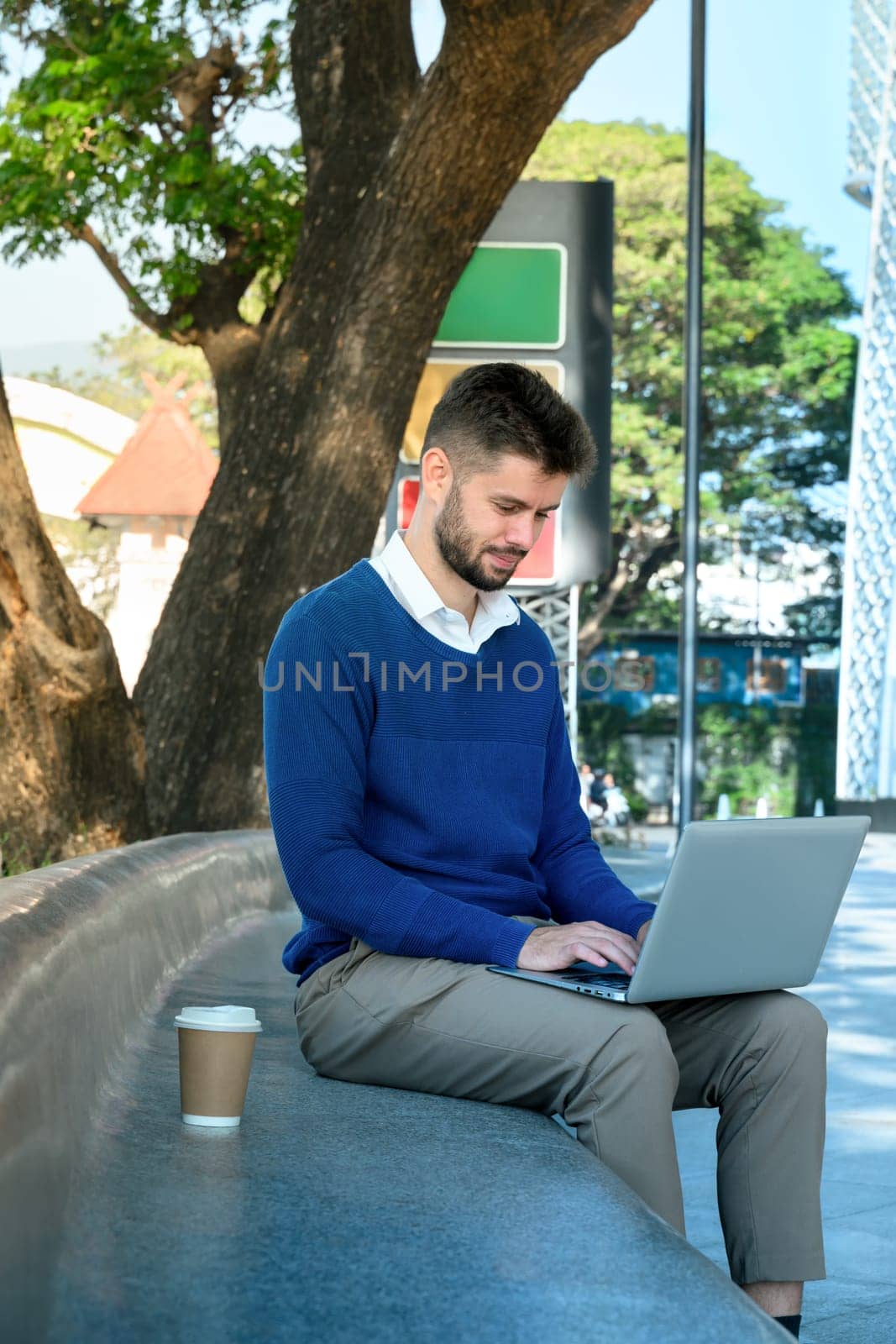 Smiling male freelancer sitting on curved bench under a tree and working on his laptop.