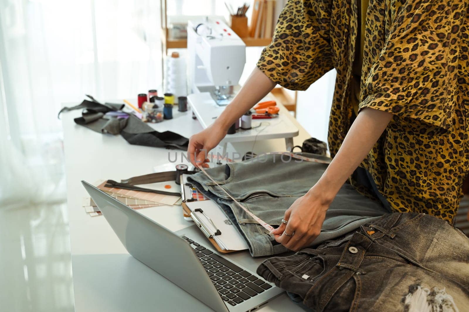Young designer measuring denim jacket with tape measure in a creative fashion studio.
