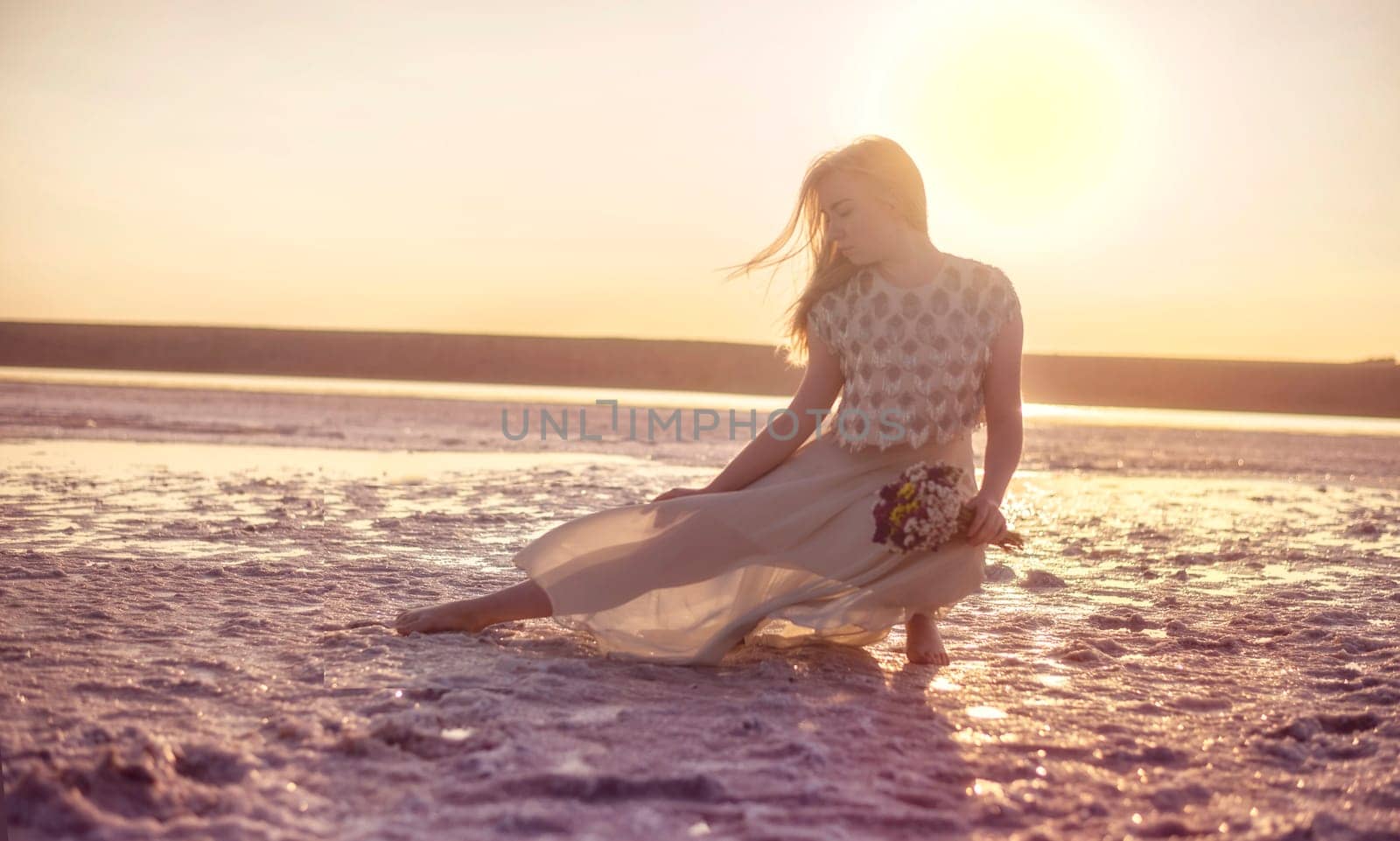 blonde girl sits in the middle of a salt lake. her dress flutters in the wind. a bright circle of the setting sun illuminates the shore covered with salt. she holds a bouquet in her hands, odessa