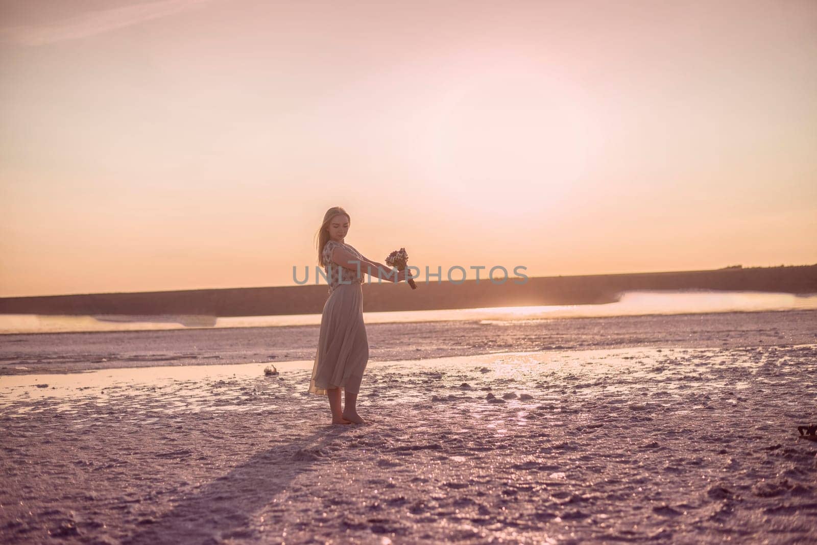 blonde girl stands in the middle of a salt lake. her dress flutters in the wind. a bright circle of the setting sun illuminates the shore covered with salt. she holds a bouquet in her hands. kuyalnik, odessa