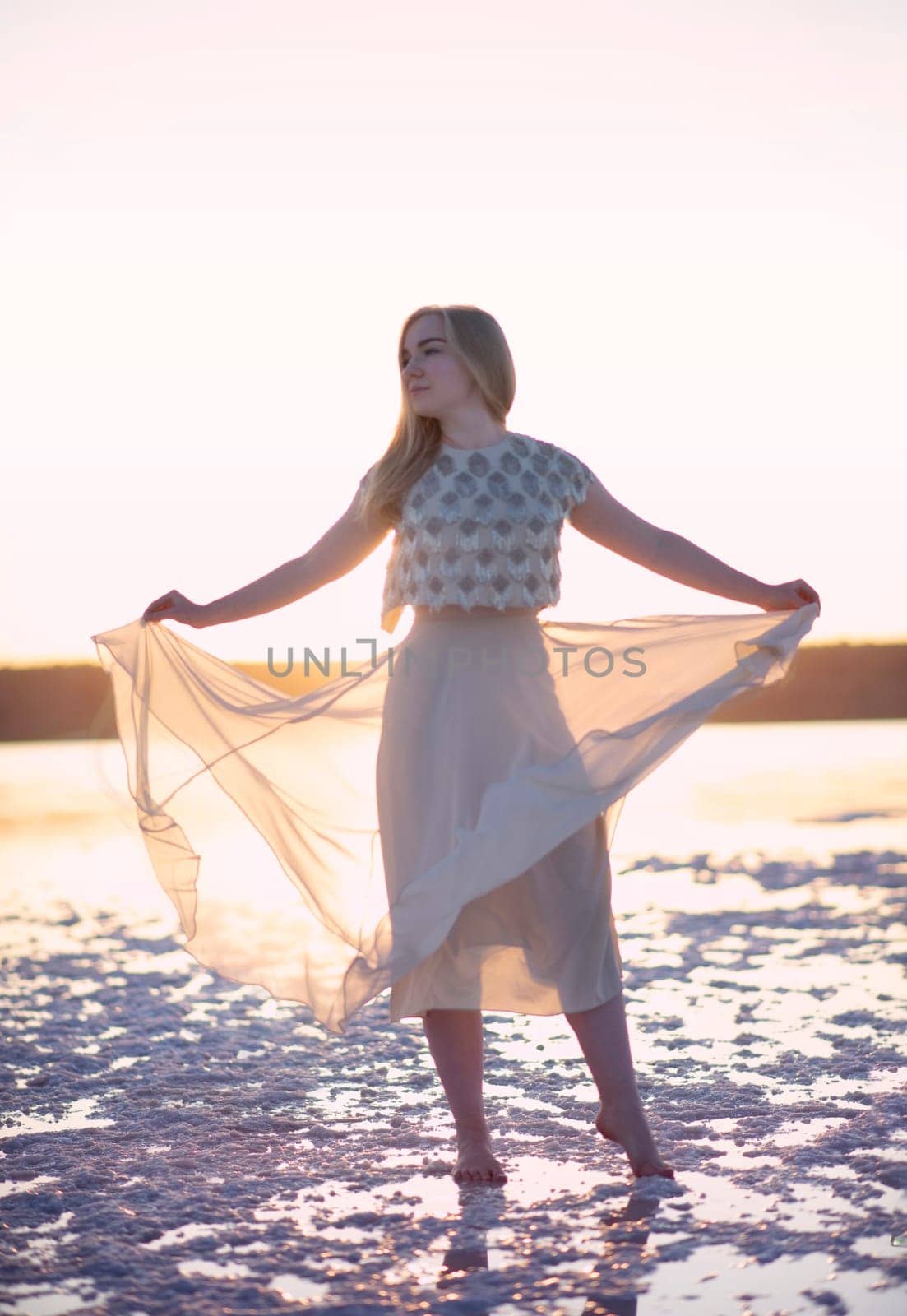 Slender blonde girl stands on the surface of sea salt at sunset. She is illuminated by the setting sun. Her chiffon dress is illuminated. Her silhouette on the horizon. Kuyalnik, Odessa, Ukraine