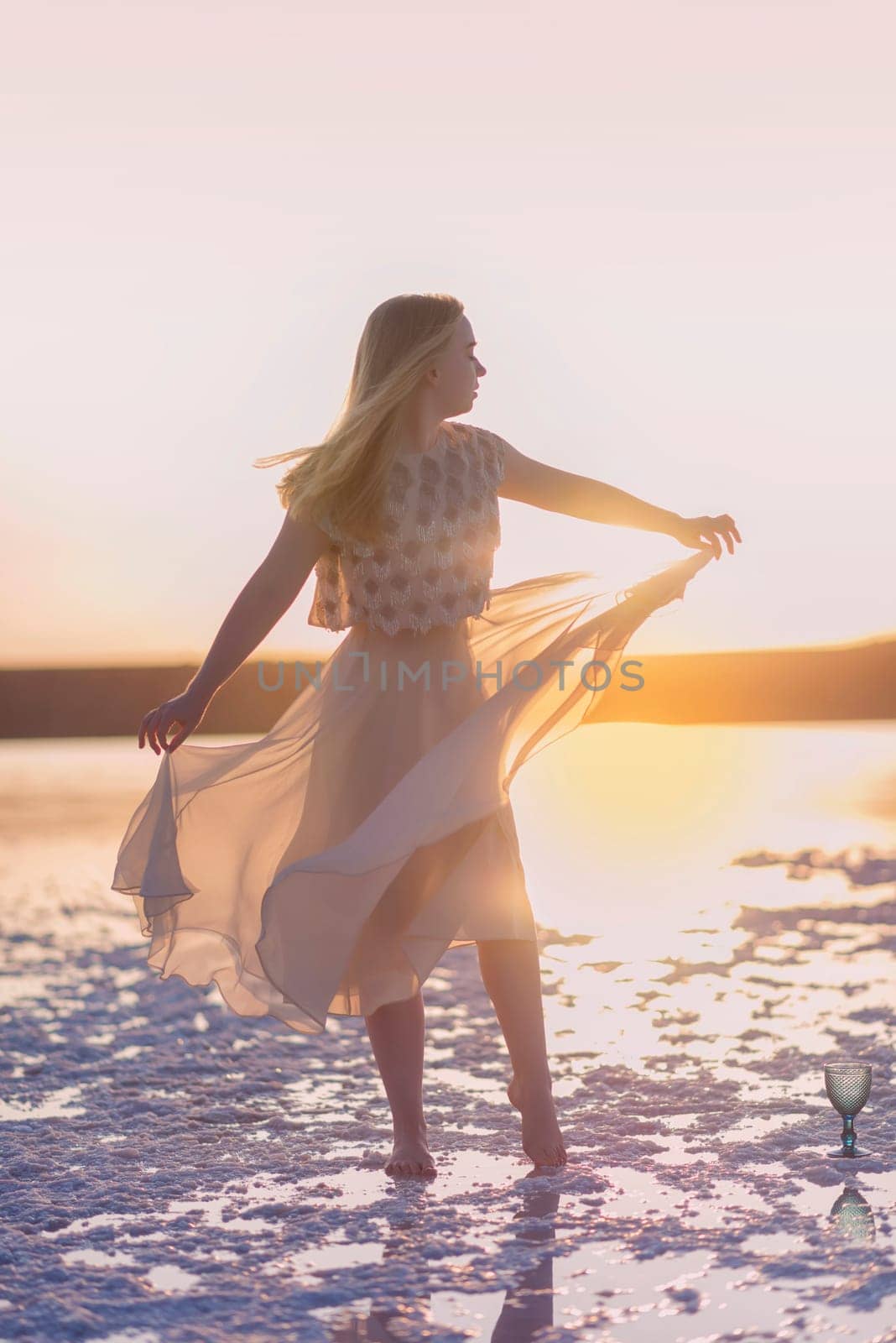 Slender girl stands on the surface of sea salt at sunset. She is illuminated by the setting sun. Her chiffon dress is illuminated. Her silhouette on the horizon. Kuyalnik, Odessa, Ukraine