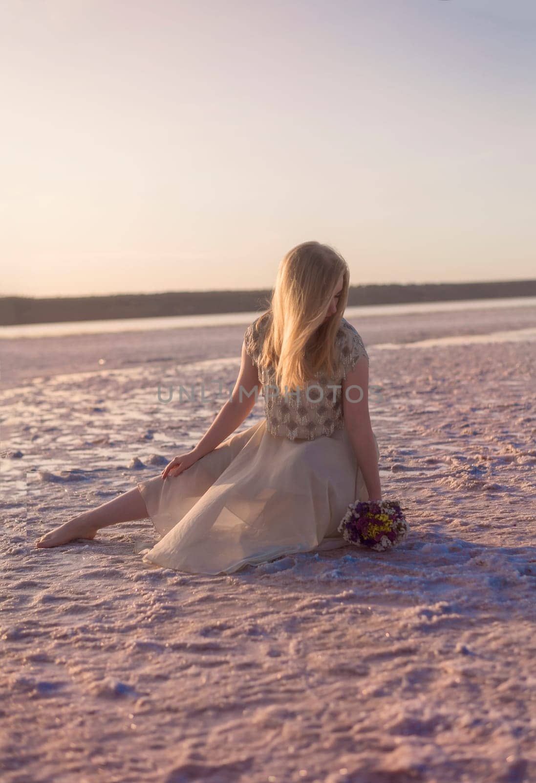 blonde girl sits in the middle of a salt lake. her dress flutters in the wind. the setting sun illuminates the shore covered with salt. kuyalnik, odessa