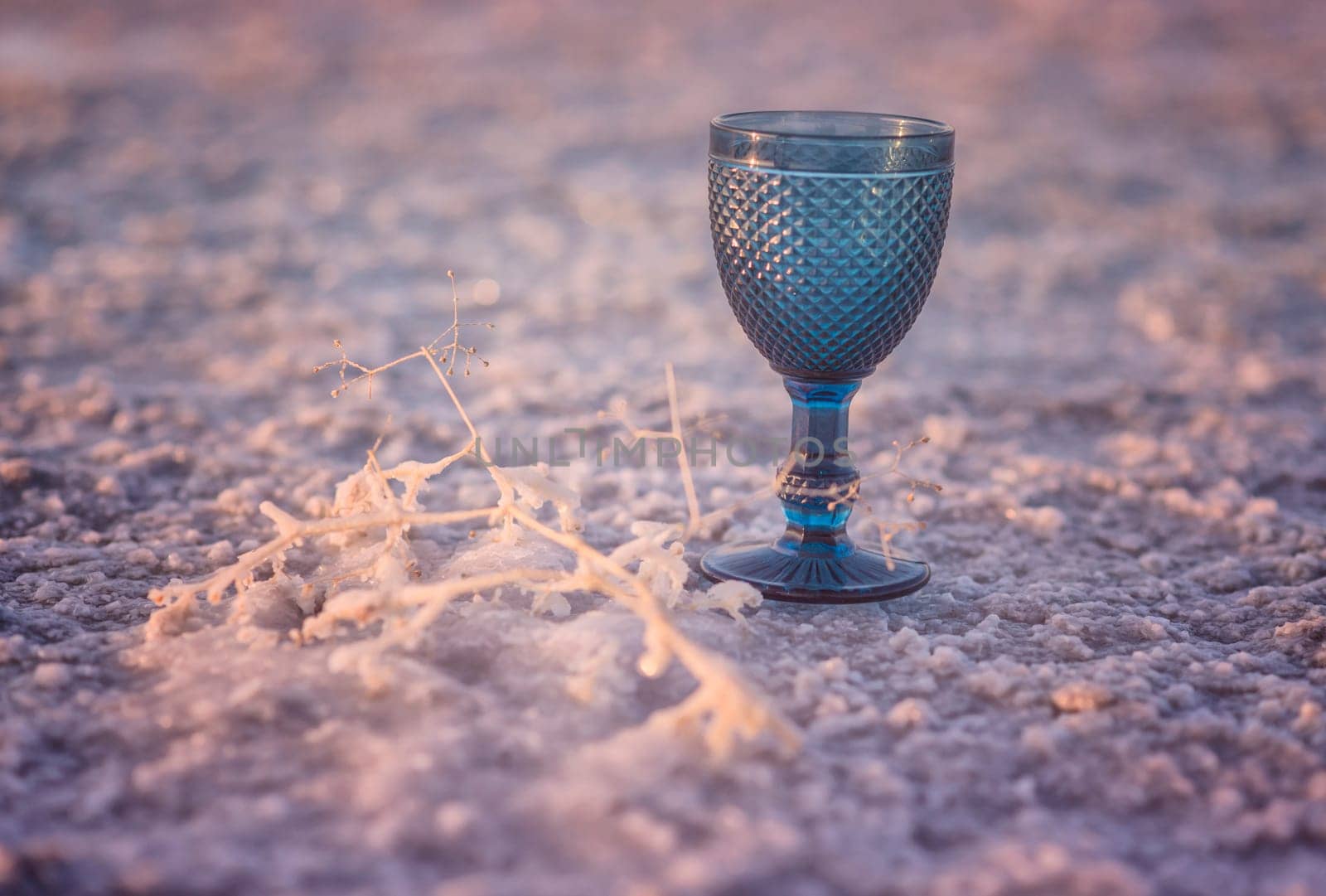 A beautiful blue glass goblet stands on a pink textured surface. Nearby is a coral branch. The earth is covered with sea salt, it shimmers in the sunA glass with a blue background. High quality photo