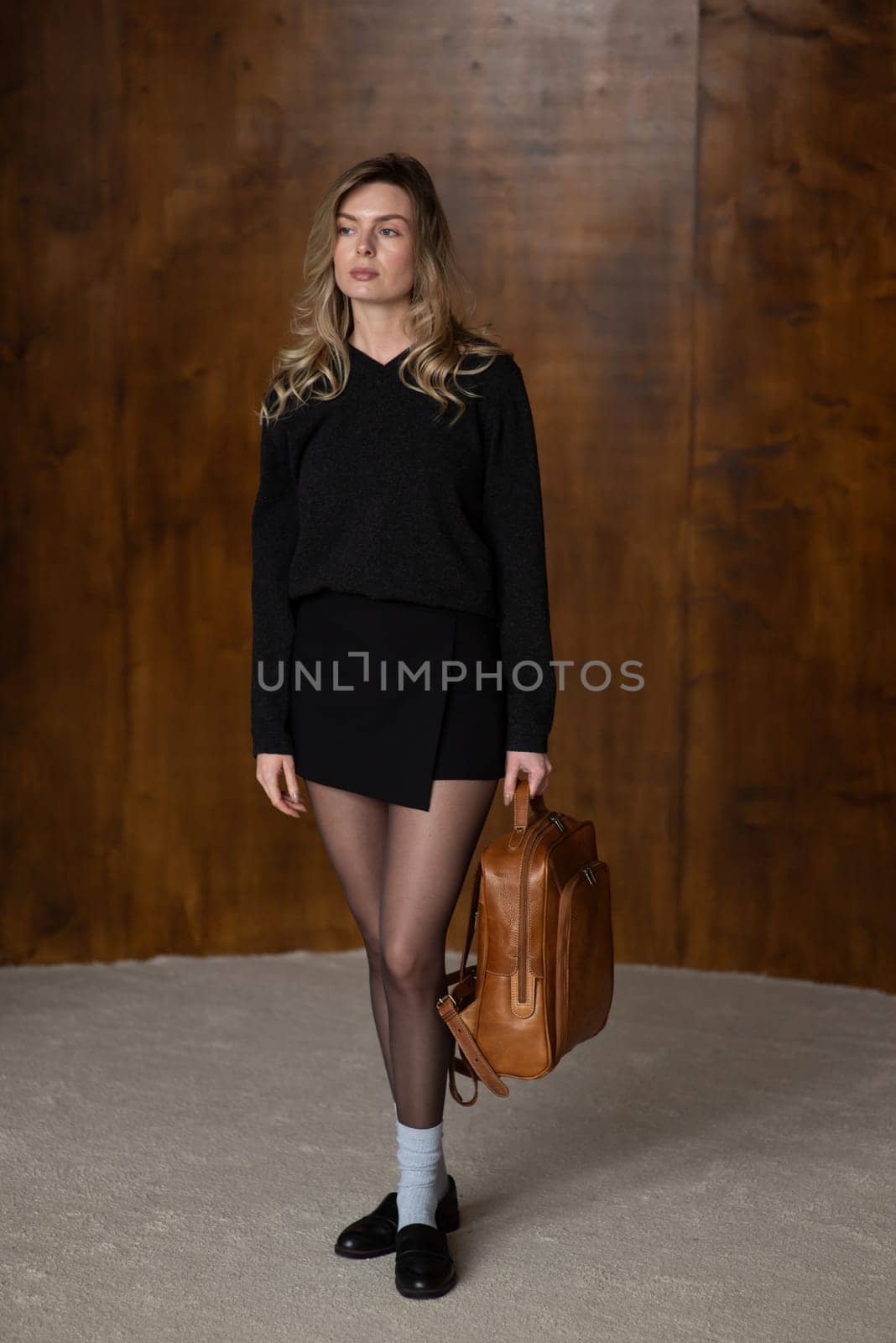 A girl in a warm sweater, a black short skirt, nylon tights, black loafers and gray socks is posing with a brown backpack