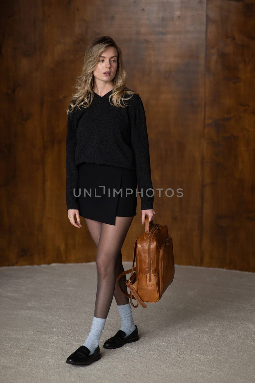 A girl in a warm sweater, a black short skirt, nylon tights, black loafers and gray socks is posing with a brown backpack