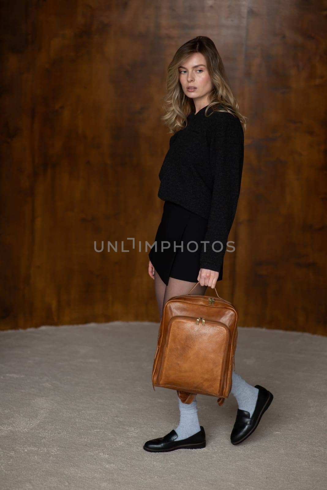 A girl in a warm sweater, a black short skirt, nylon tights, black loafers and gray socks is posing with a brown backpack