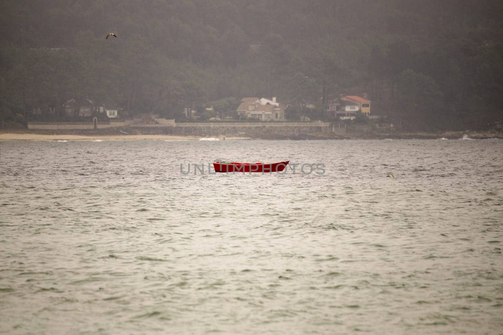 A vivid red boat gently floats on the tranquil ocean, surrounded by fog, creating a serene atmosphere reflecting peace and solitude amid visible coastal homes.