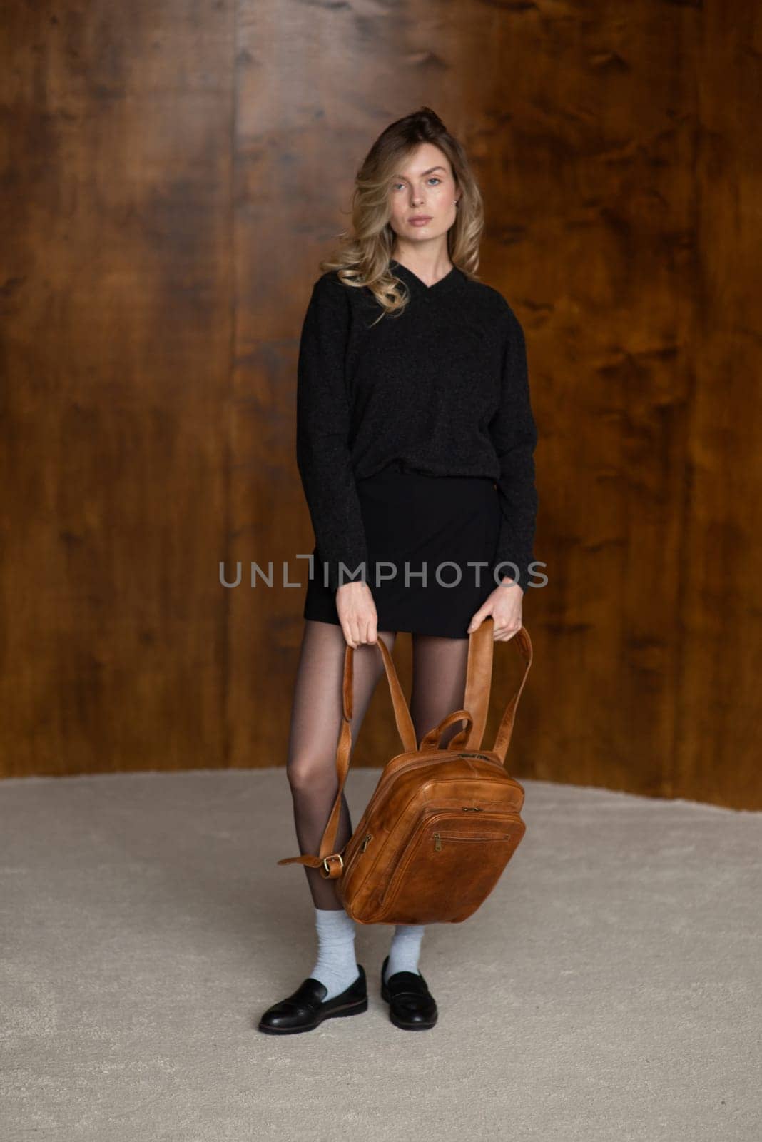 A girl in a warm sweater, a black short skirt, nylon tights, black loafers and gray socks is posing with a brown backpack