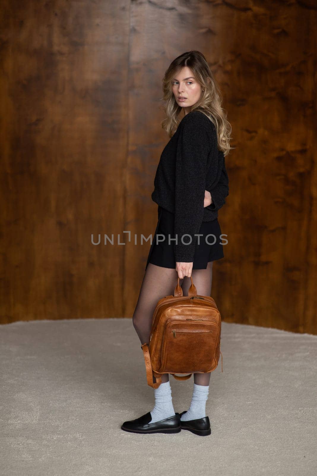 A girl in a warm sweater, a black short skirt, nylon tights, black loafers and gray socks is posing with a brown backpack