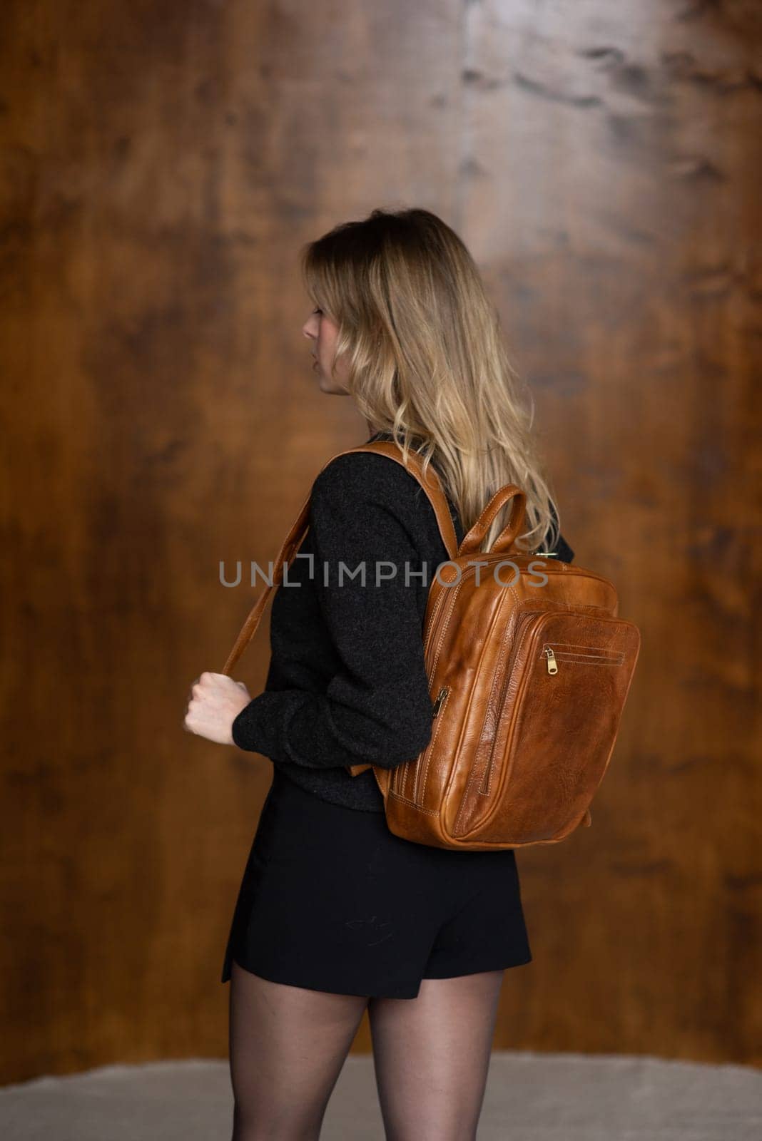 A girl in a warm sweater, a black short skirt and nylon tights is posing with a light brown backpack