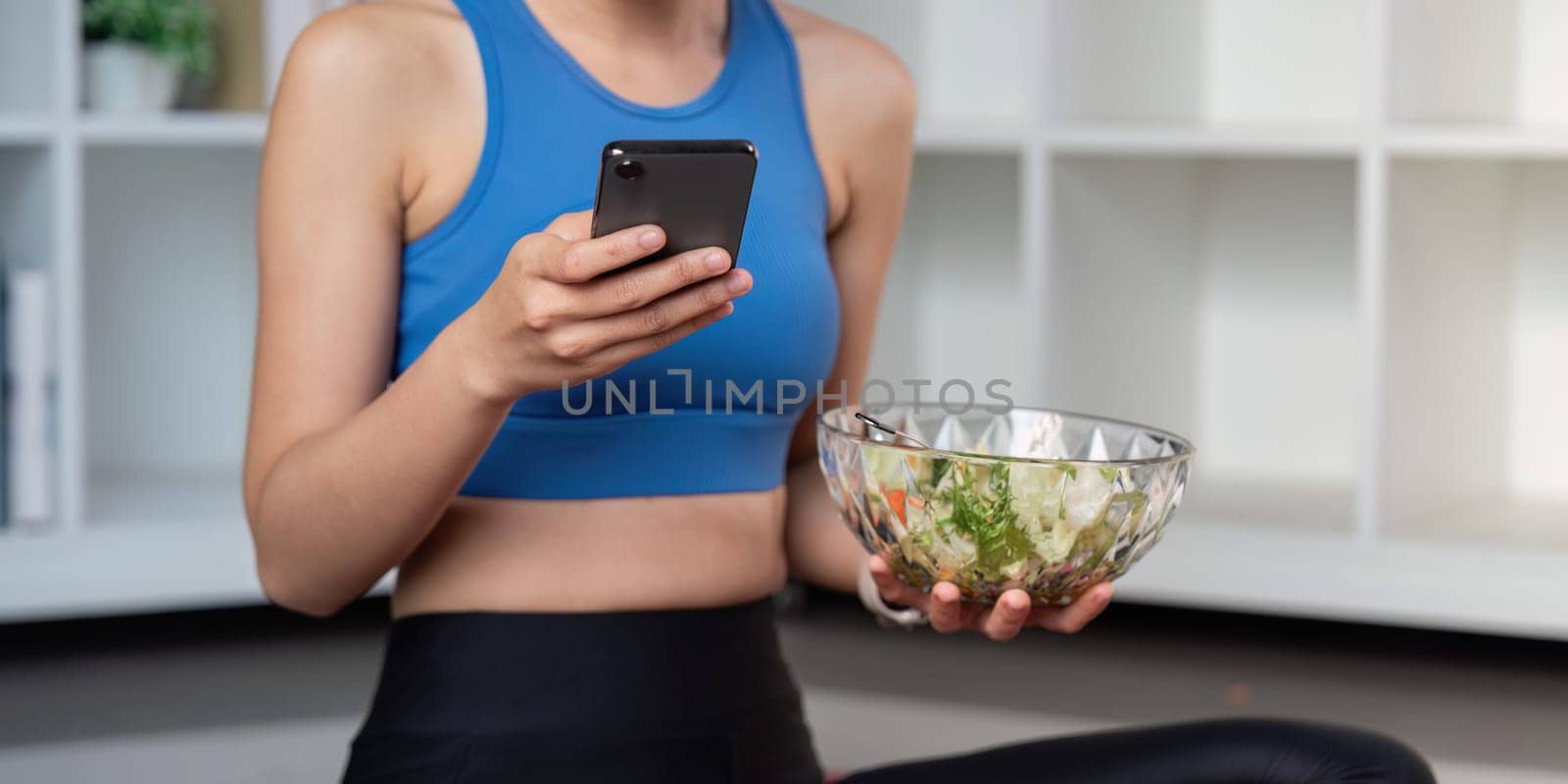 A fit woman in activewear checks her smartphone while holding a bowl of fresh salad, embodying a healthy lifestyle.