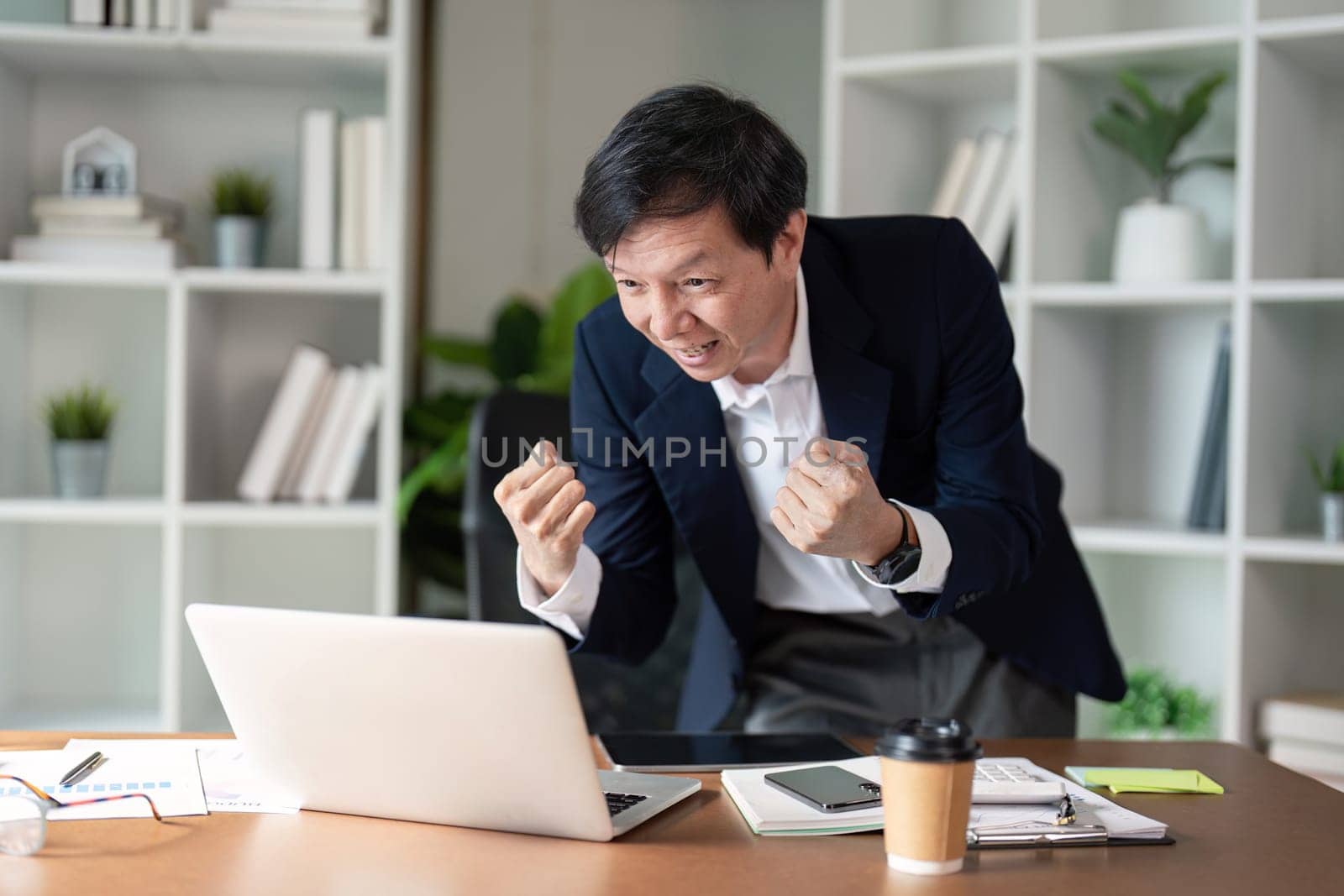 An elderly businessman expresses excitement and triumph after achieving a milestone while working on his laptop.