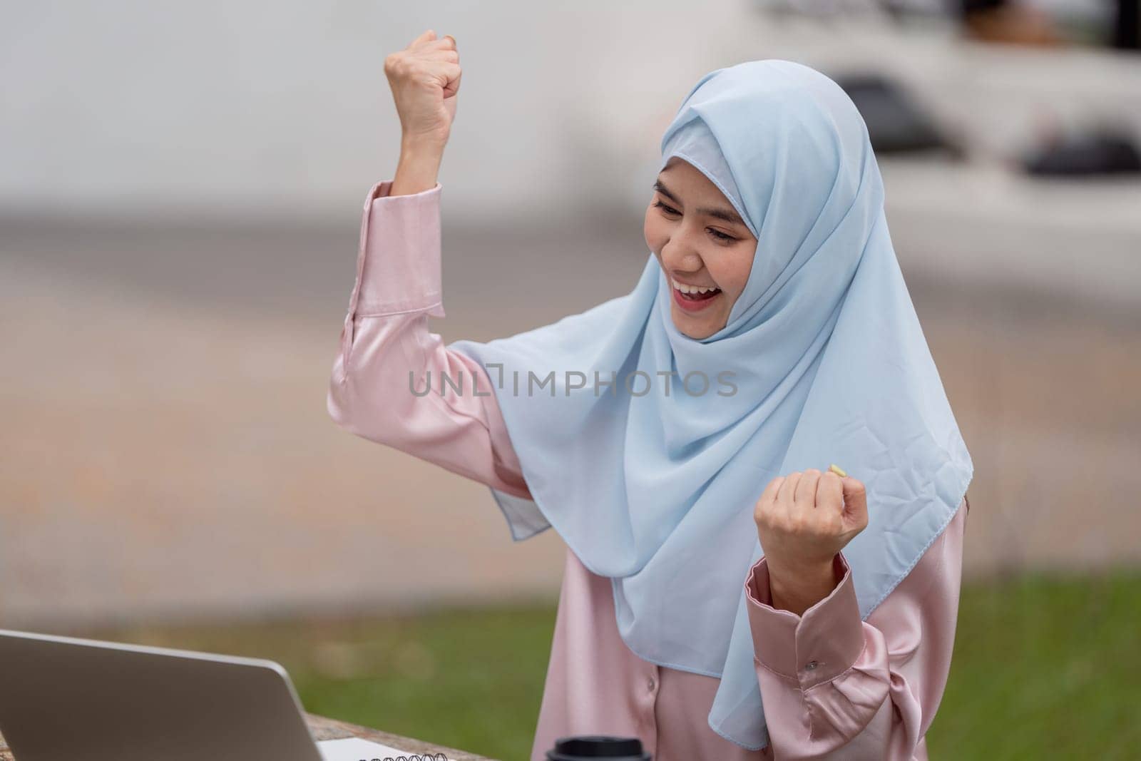 A happy Muslim woman in a hijab celebrates her achievements while working on a laptop in an outdoor setting, showcasing empowerment.