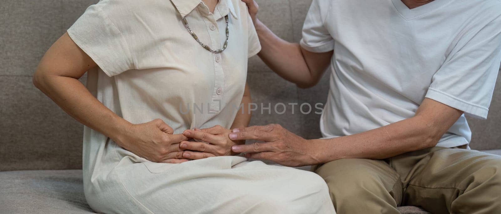 An elderly couple on a couch, one partner holding their abdomen in discomfort while the other offers support and care.