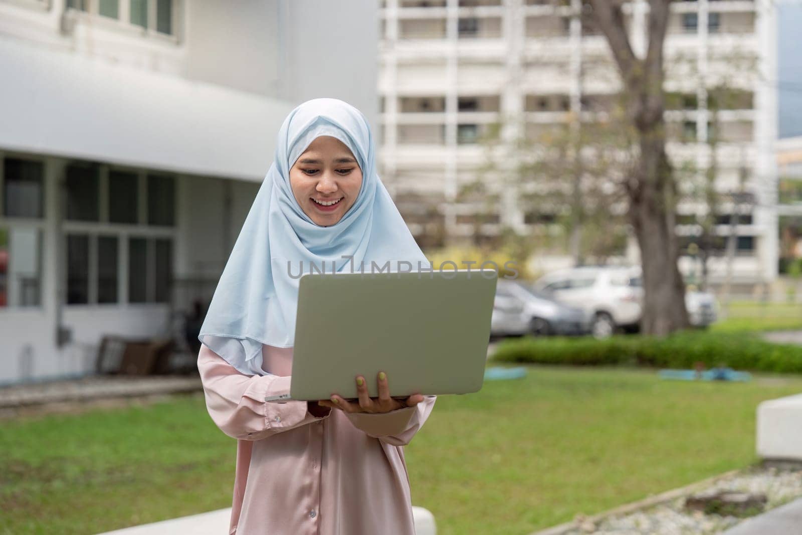 A joyful Muslim woman in a hijab smiles while using her laptop outdoors, showcasing positivity in her business endeavors.