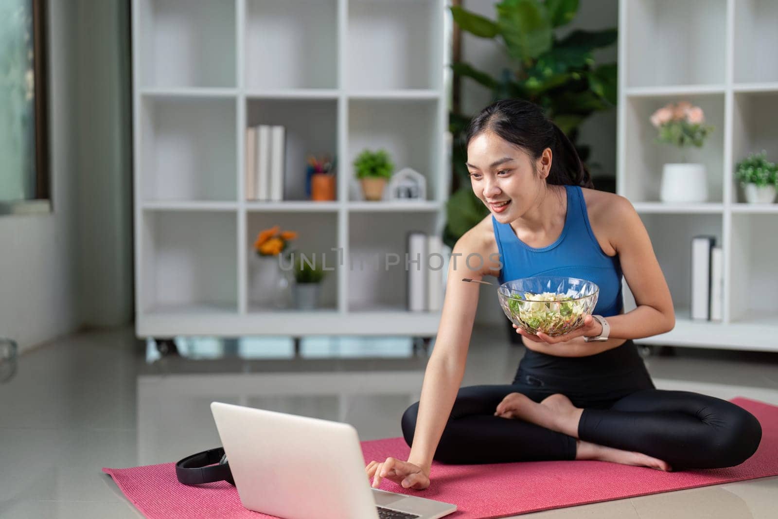 A woman in activewear smiles as she uses her laptop while eating a fresh salad, blending nutrition with technology.