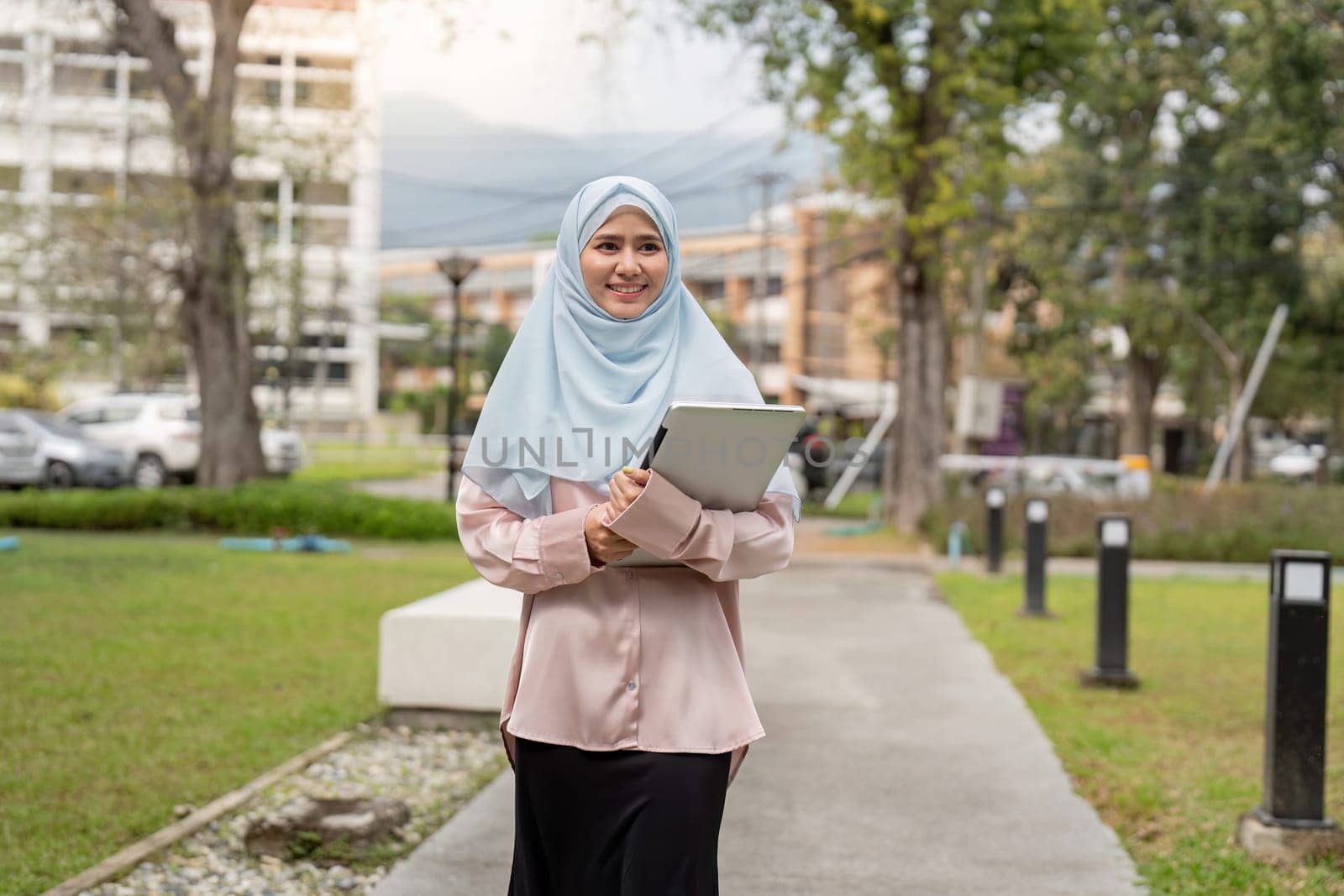 A cheerful Muslim woman in a hijab walks through a park, holding a laptop and exuding confidence and professionalism in her business role.