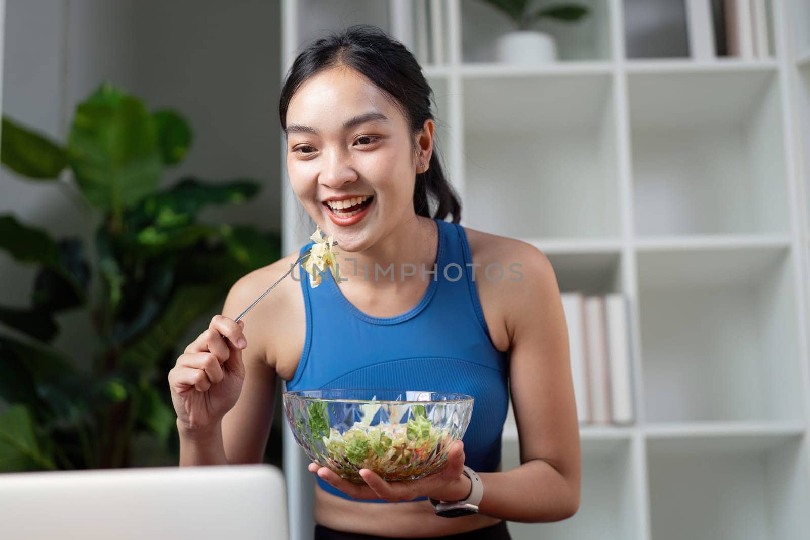 A happy woman laughs while enjoying a salad and engaging with her laptop, showcasing a blend of nutrition and technology.