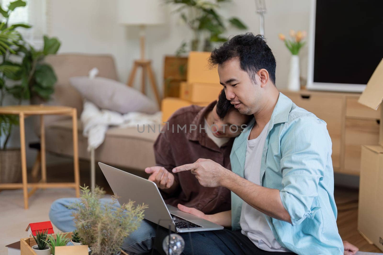 A gay couple sits together, working on a laptop while unpacking boxes in their new home, showcasing teamwork and love.