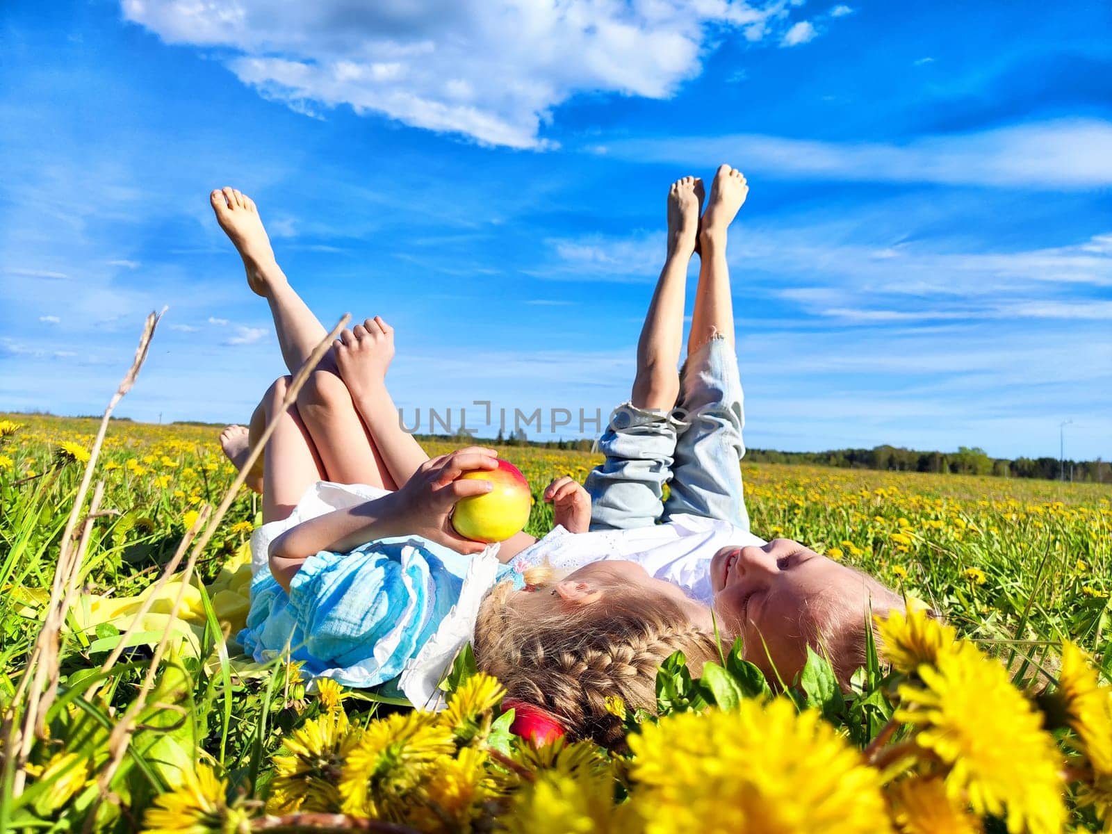 Two children lie on their backs in a vibrant dandelion field, enjoying fresh air and a bright sunny day while holding an apple.