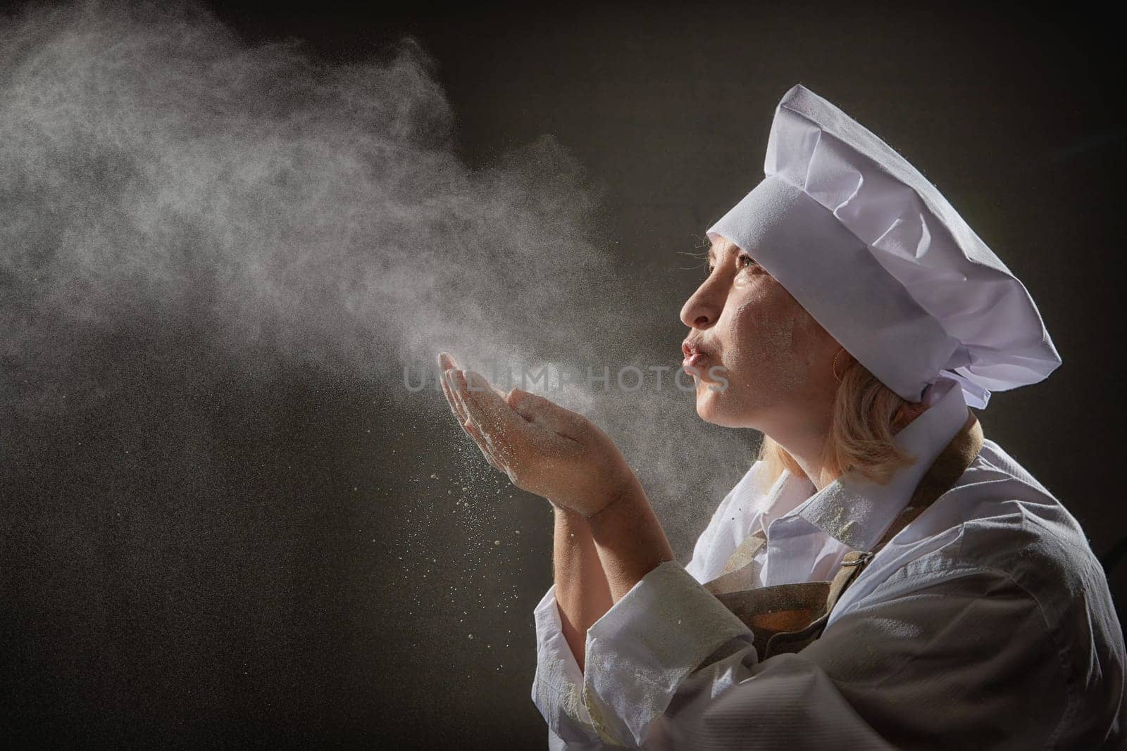An elderly woman dressed as a cook joyfully blows flour while enjoying a lighthearted kitchen photoshoot with whimsical expressions.