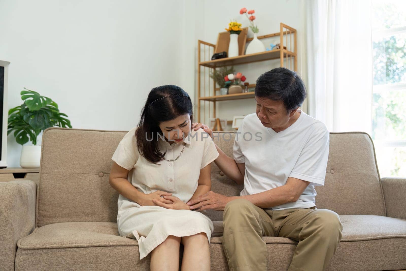 An elderly couple sitting on a couch, with one partner expressing discomfort while the other provides comfort and care.