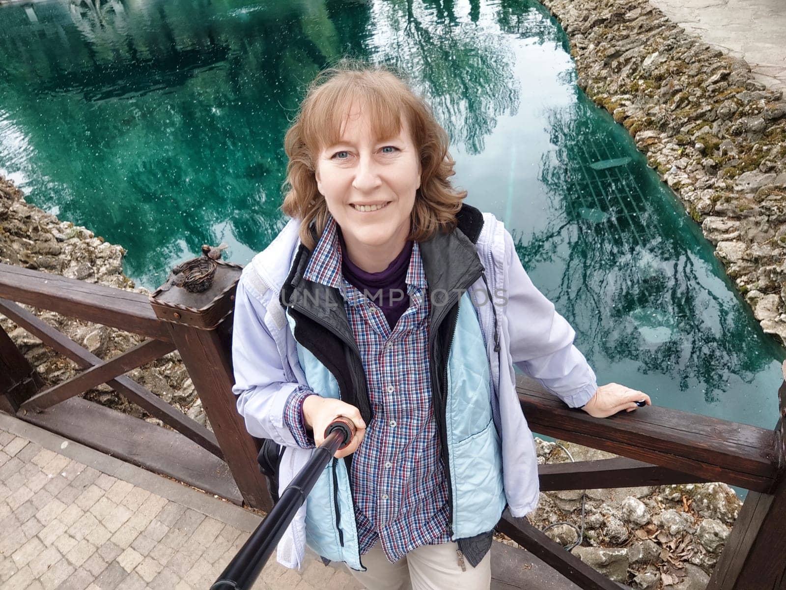 A middle-aged woman with blonde hair captures selfies in a beautiful park during her trip, surrounded by lush nature.