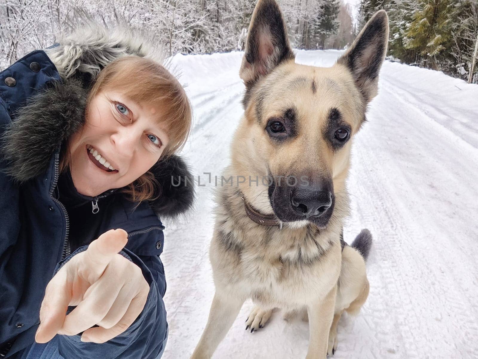 A middle-aged woman enjoys a winter camping trip, taking a cheerful selfie with her German shepherd dog in the snow.