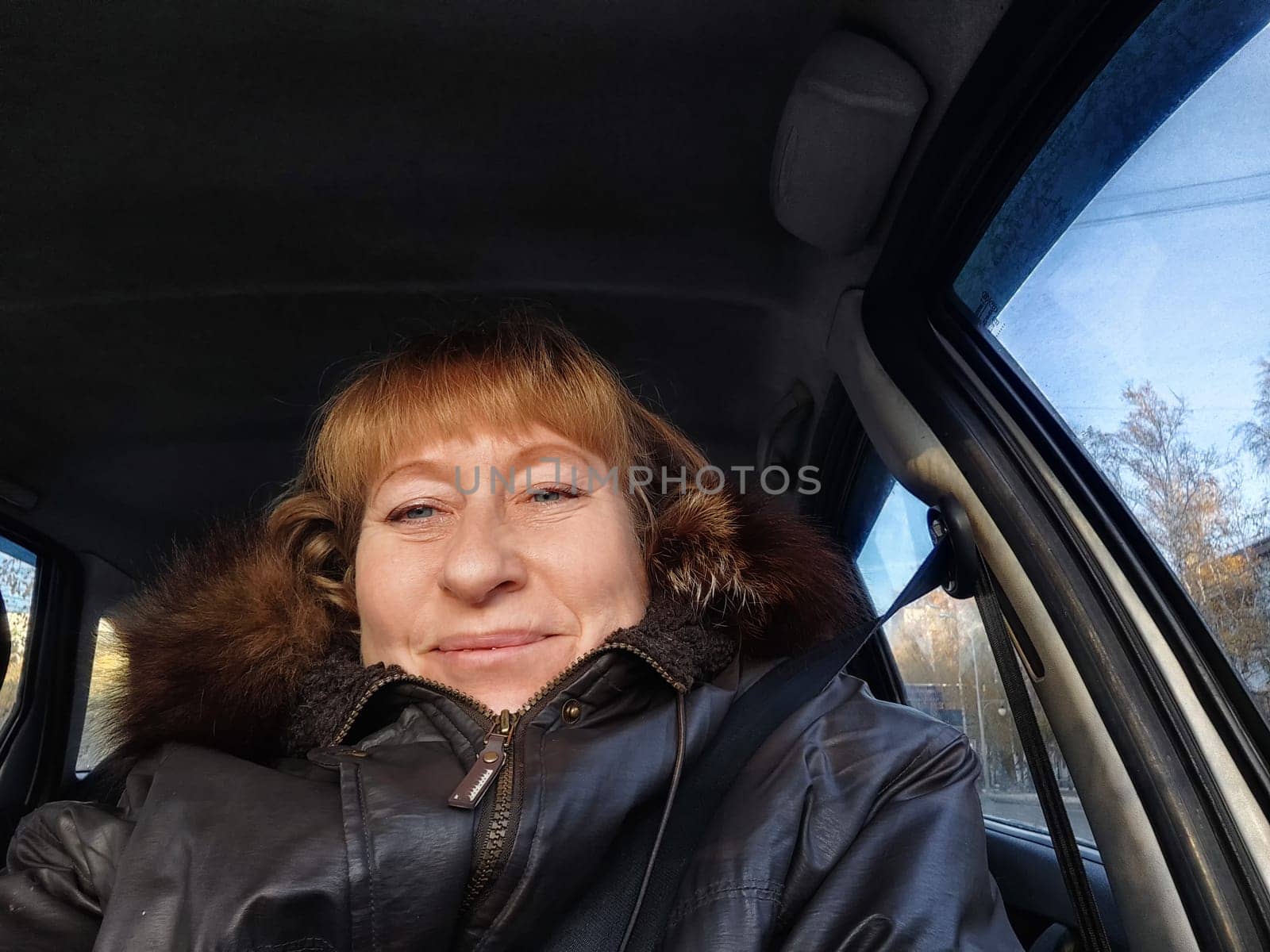 A middle-aged woman smiles while taking a selfie from the driver's seat during her adventurous road trip through scenic landscapes.