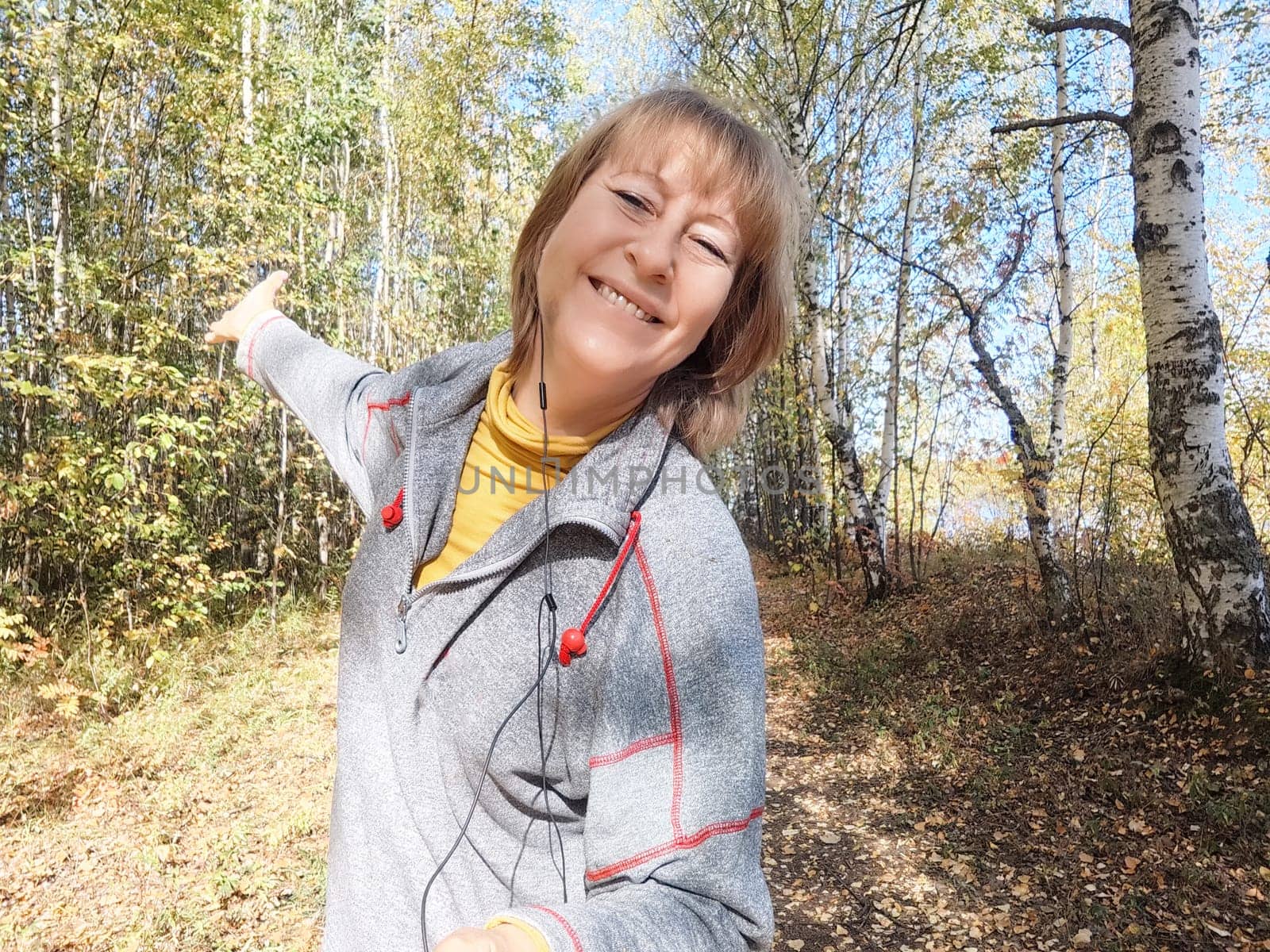 Blonde woman on a nature adventure, smiling amidst autumn leaves, celebrating the beauty of the outdoors in a serene landscape.