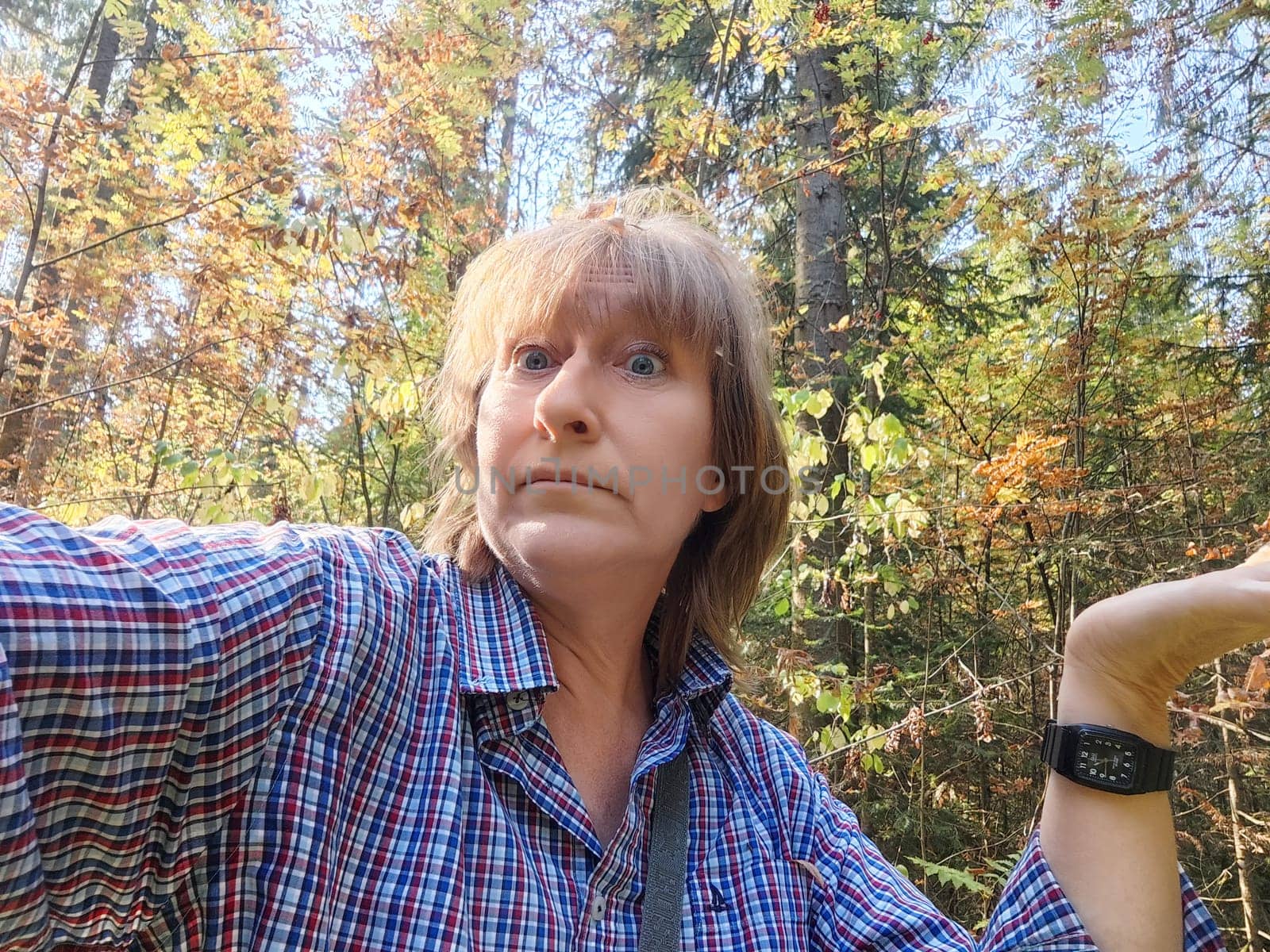 A middle-aged woman playfully makes funny faces while hiking through a beautiful forest filled with colorful foliage.