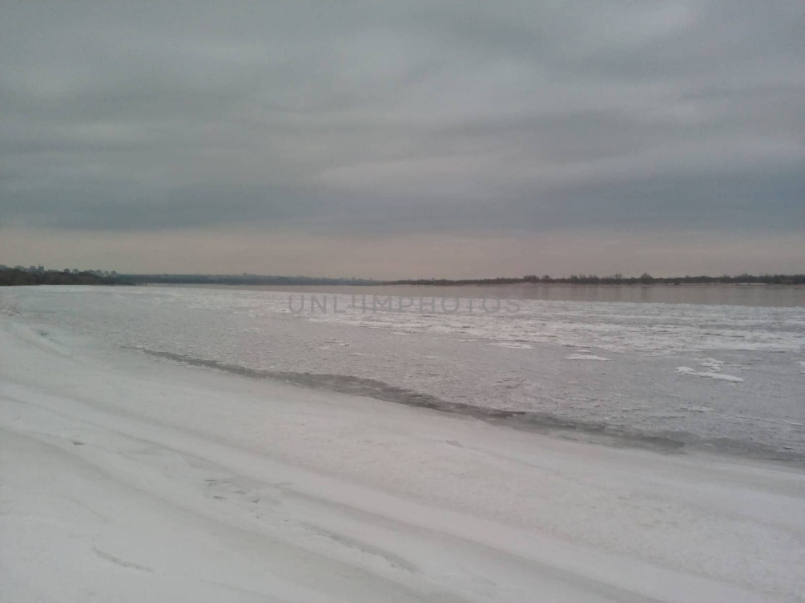 Amidst the calm of winter, the riverbank transforms into a serene landscape where snow covers the ground, evoking a sense of quiet reflection. The soft hues of the cloudy sky meet the horizon, creating a stunning juxtaposition of water and white.