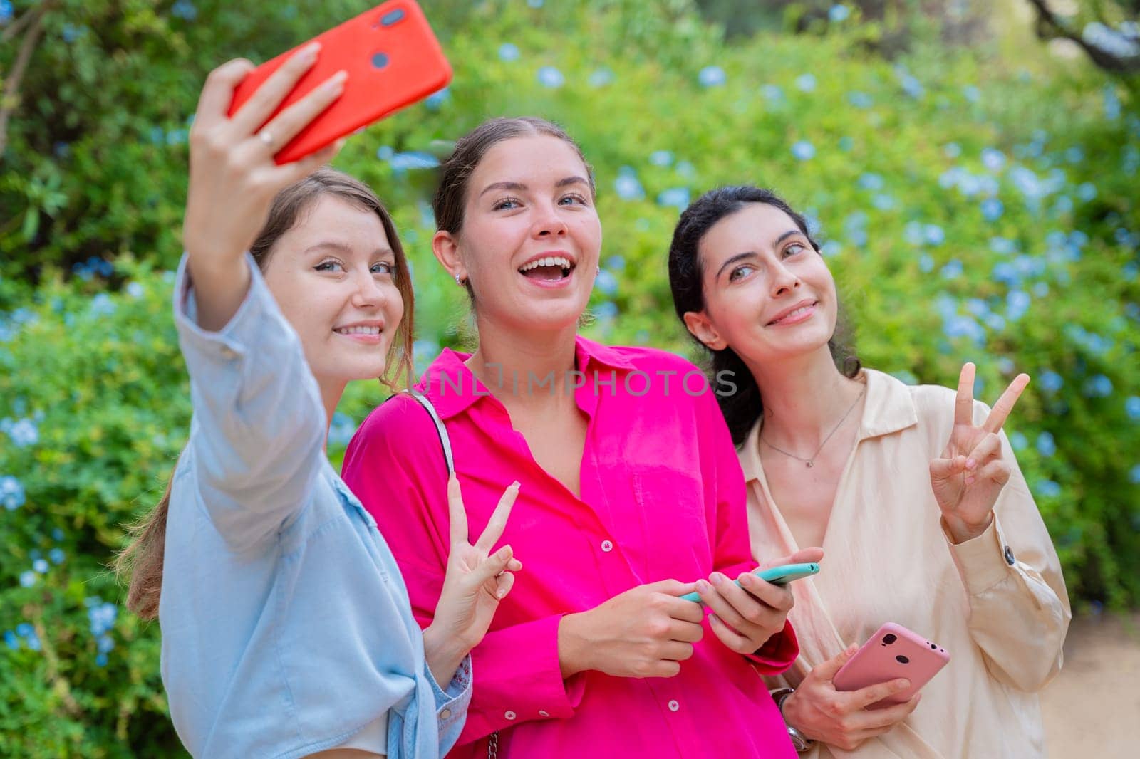 Three smiling women posing for a selfie in a lush green park. They embrace each other, expressing happiness and friendship. The vibrant colors and natural background create a joyful atmosphere.
