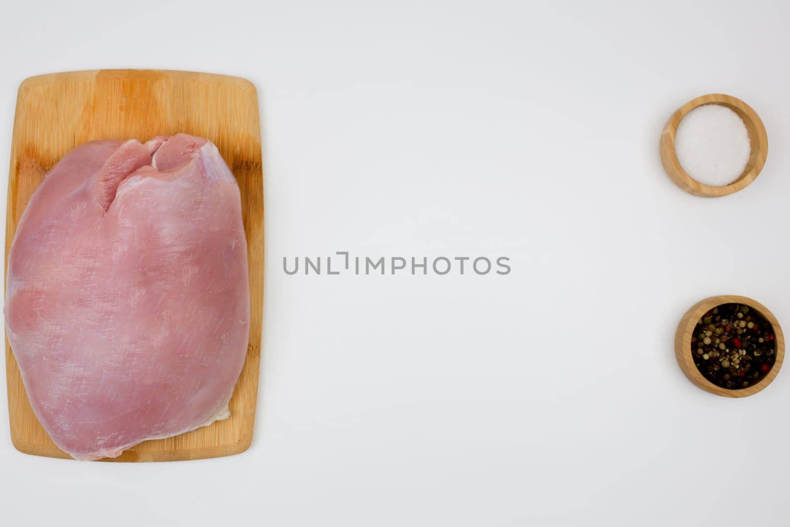 On the left is a piece of turkey fillet brisket, which lies on a wooden cutting board on a white background, on the right are seasonings in wooden bowls with space for text in the center. The concept of healthy eating, dietary and natural products, cooking. High quality photo
