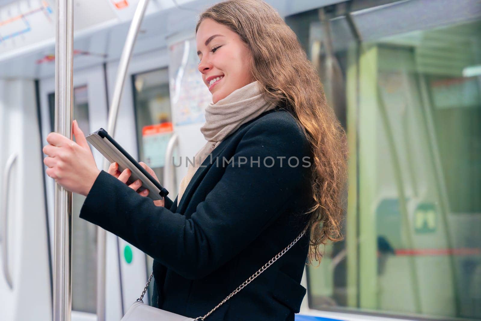 Stylish young woman in a black coat and beige scarf reading on an e-reader while standing inside a modern subway train. She balances while holding onto a pole during her commute.