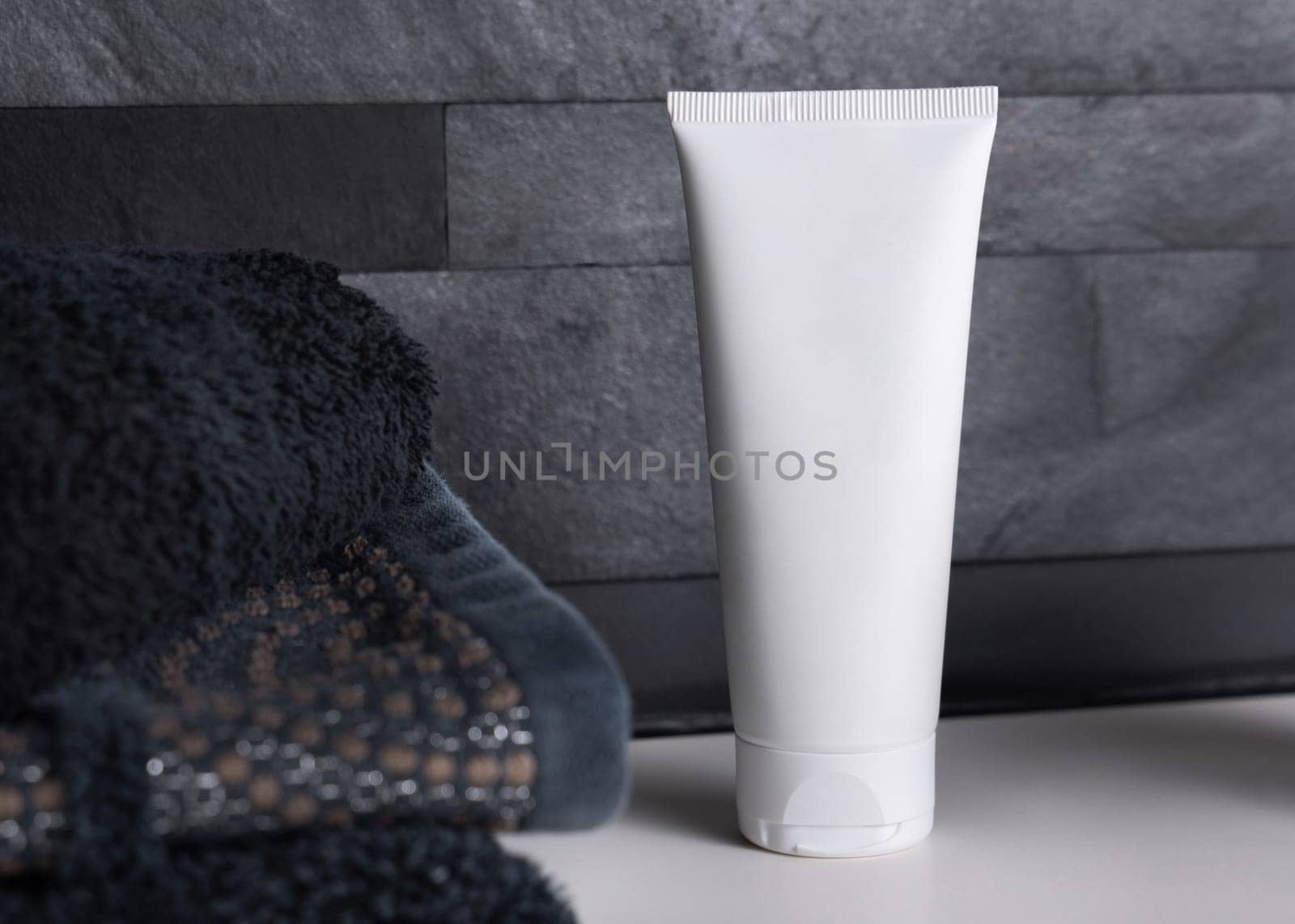 White cream squeeze tube near black folded towels on basin in dark grey masculine bathroom close up, packaging mockup. Natural plant unbranded cosmetics with copy space. Spa and wellness concept