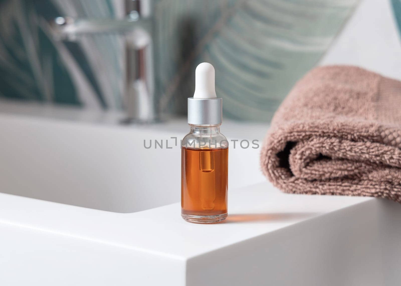 Glass dropper bottle with pipette filled with orange liquid near brown towels and faucet on basin in green tropical bathroom close up. Natural cosmetics - serum or essence. Spa concept