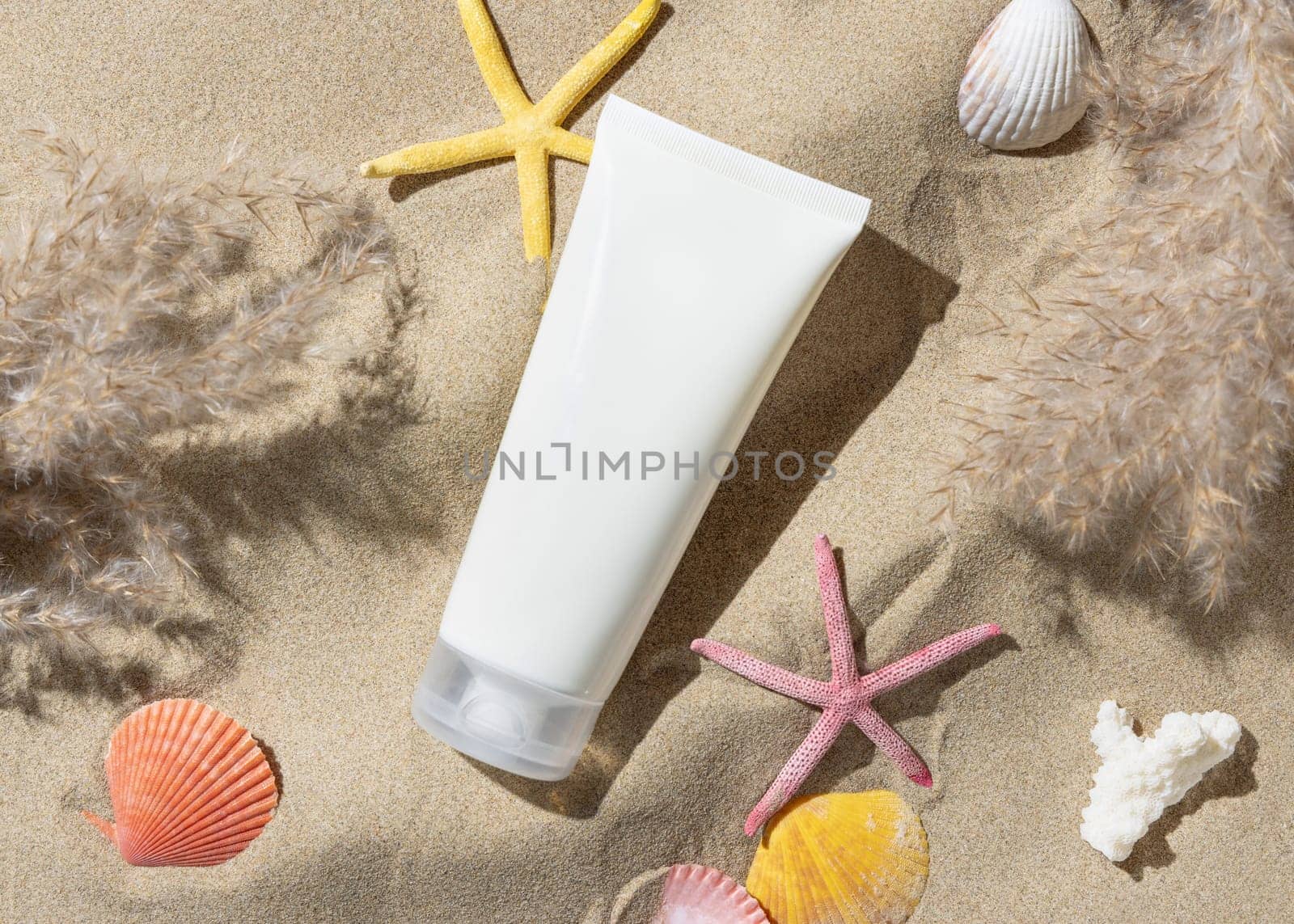White blank cream tube, starfishes and seashells on a sandy beach top view, hard shadow. Sunscreen cosmetics mockup. Summer vacation with spf uv-protected sunbathing