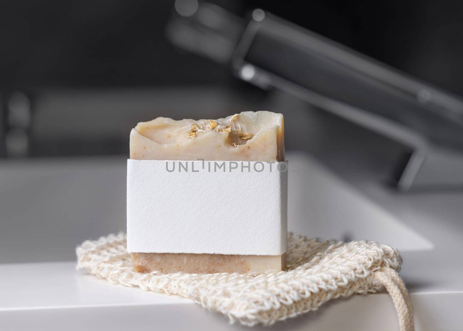 Beige handmade soap bar with blank label on sisal soap saver bag near modern faucet on basin in dark grey masculine bathroom close up, mockup. Natural herbal product for daily hygiene and bodycare