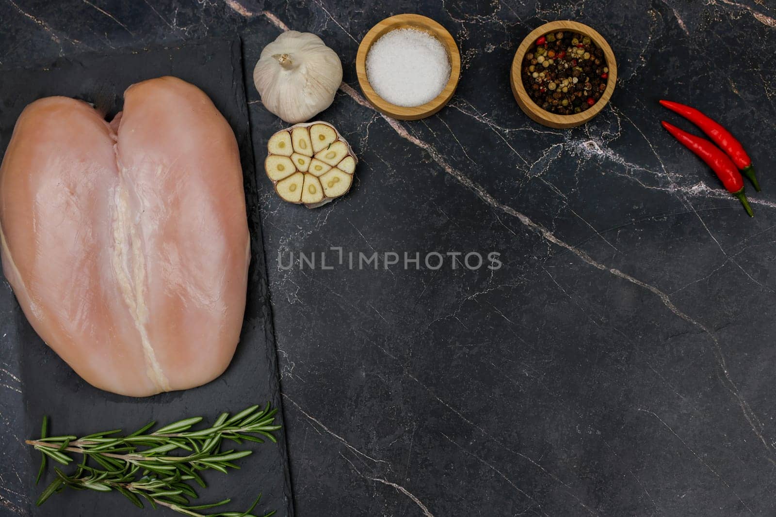 Chicken breast fillet lies on a black board on a dark background with a sprig of rosemary next to it, garlic, red pepper and spices and salt on top in wooden bowls with space for text. The concept of proper and healthy nutrition, dietary dishes and recipes. High quality photo