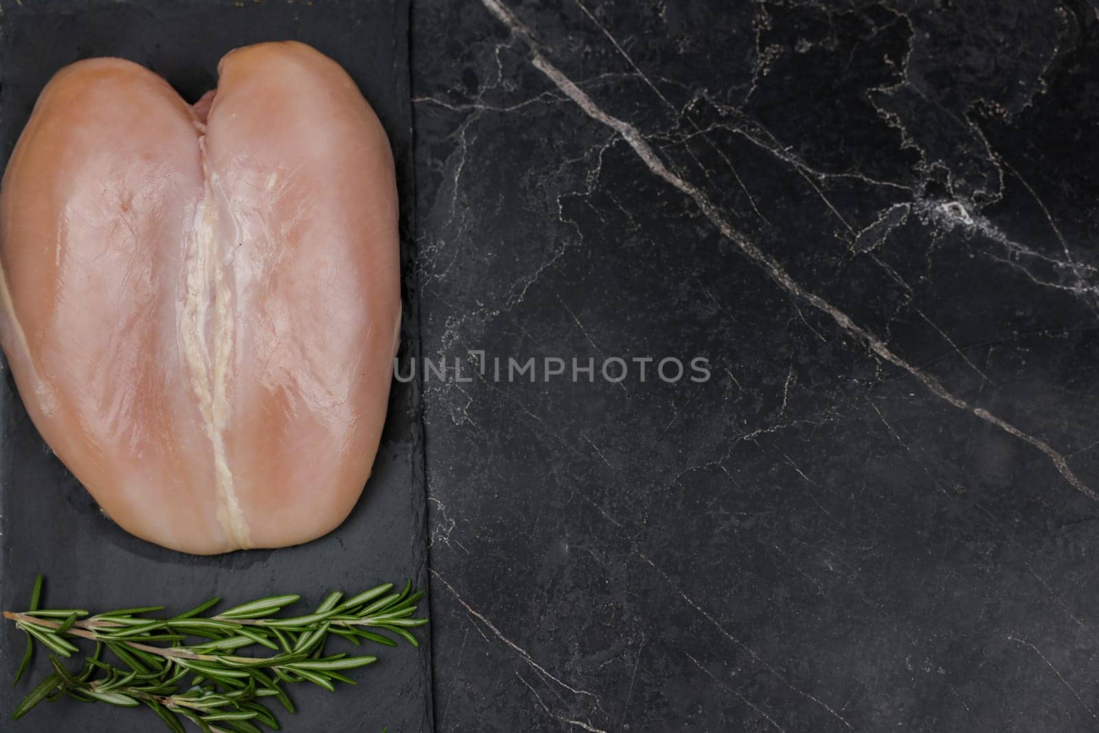Chicken breast fillet lying on a black board on a dark background with a sprig of rosemary on a dark background with space for text. The concept of proper and healthy nutrition, dietary dishes and recipes. High quality photo